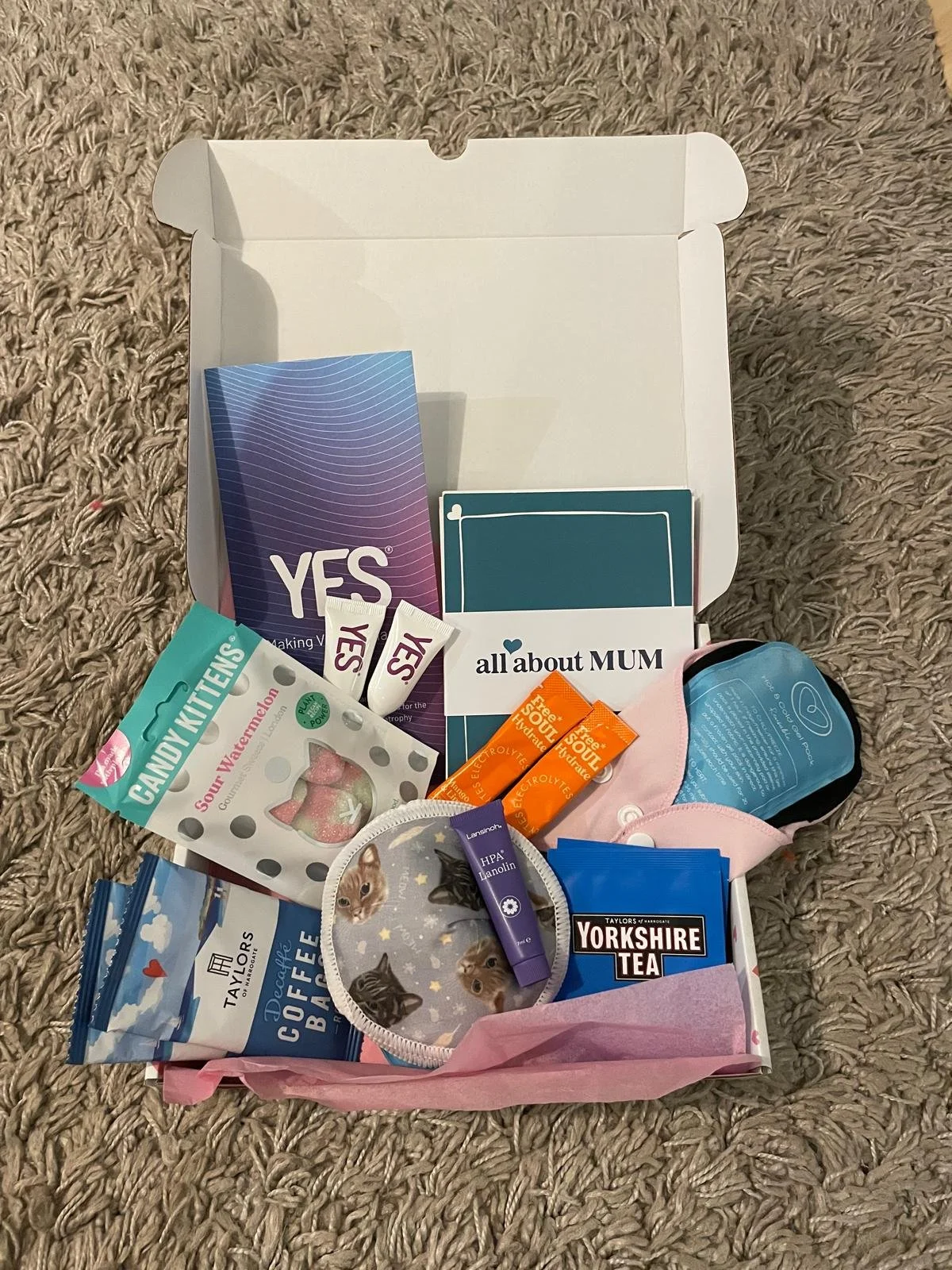 Open gift box with various items inside including tea, snacks, vaginal moisturiser, an ice pack for the perineum. A perineal ice pack. A perineal heat pack for comfort. Breast pads included. All about Mums cards to give advice when a mum is newly pos