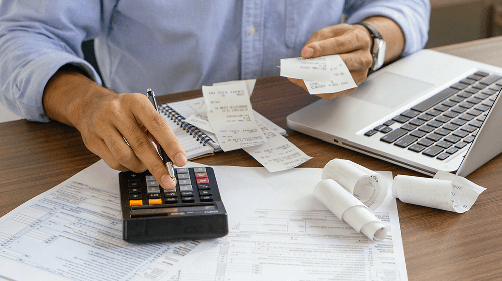 Person calculating expenses with receipts, calculator, and laptop on desk.
