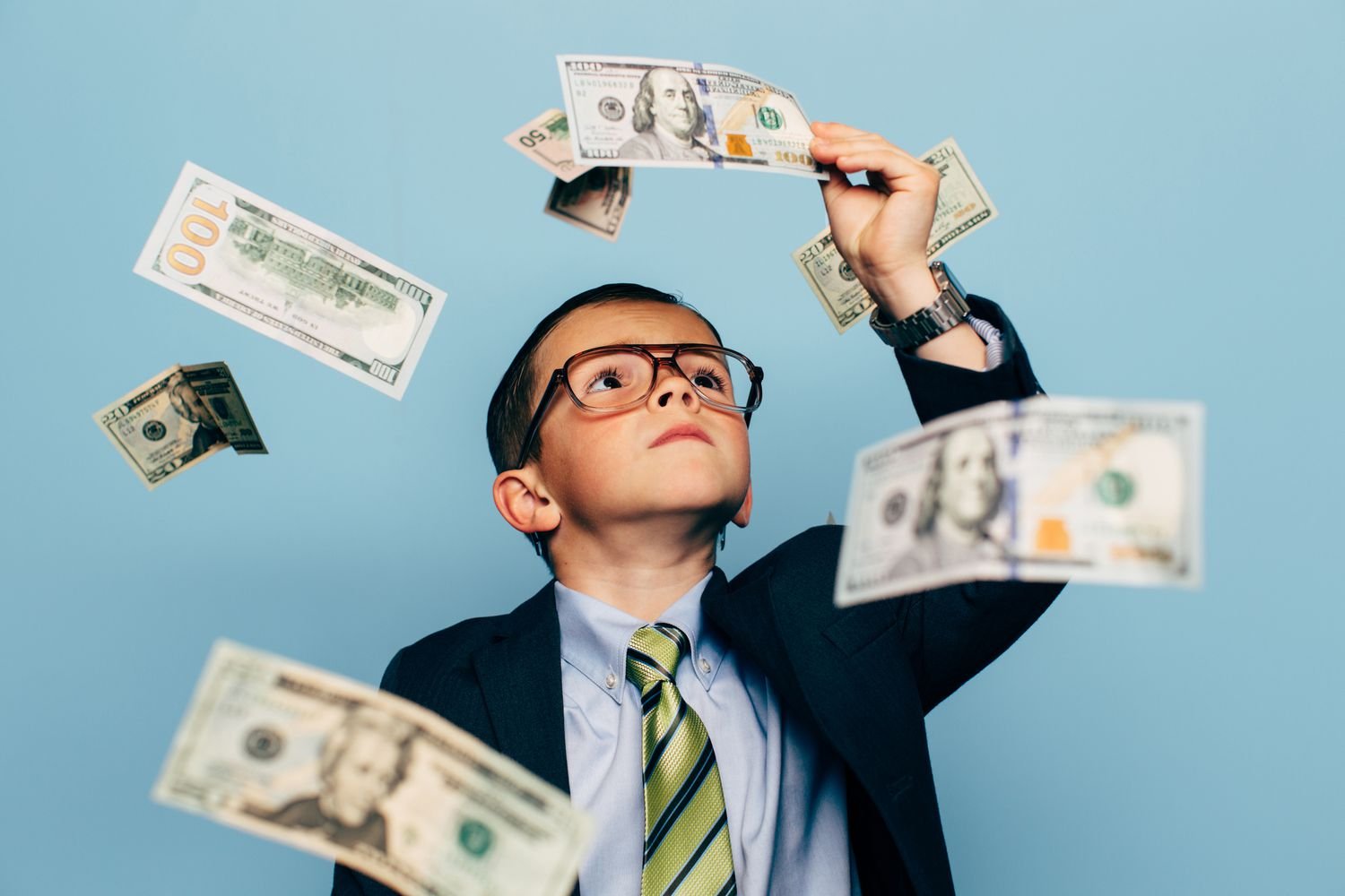A young boy in a business suit and glasses is throwing dollar bills into the air.