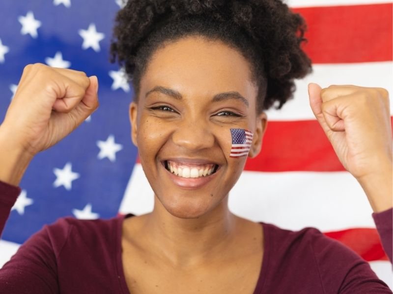 A smiling woman with an American flag sticker on her cheek, showing strength with her clenched fists, standing in front of an American flag background.