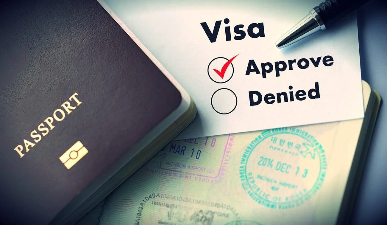 A passport, a pen, and a sheet of paper with a checkmark next to the word 'Approve' for a visa application, with visible stamps on the paper.