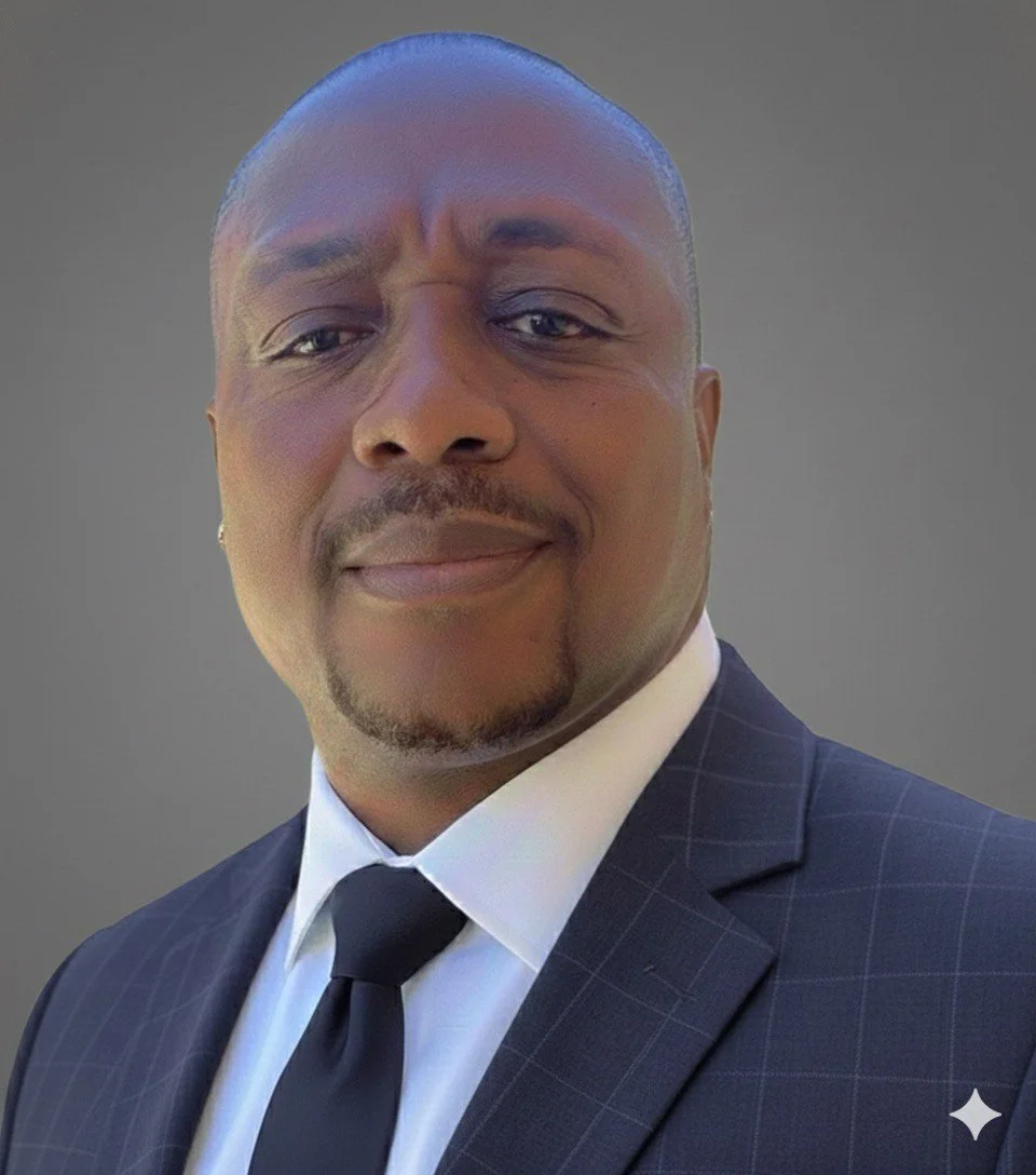 Close-up of a smiling man in a dark checked suit, white shirt, and black tie, against a plain gray background.