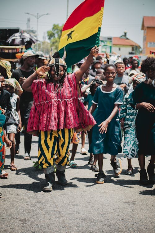 A group of people, mostly children, participating in a street celebration or rally, with a woman holding a Ghanaian flag.