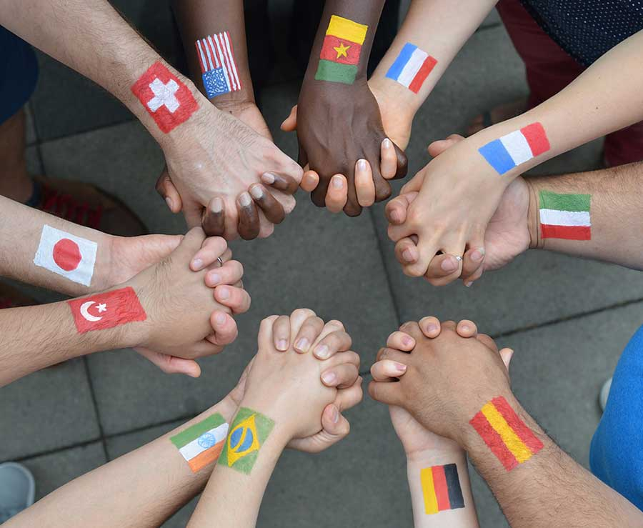 Group of diverse people holding hands with flags painted on their arms, representing different countries including Switzerland, Japan, Turkey, United States, Ghana, France, Italy, Brazil, Germany, and Poland.
