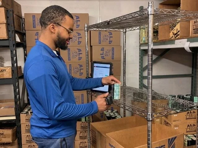 Man in blue jacket holding a barcode scanner in warehouse with shelves and boxes.