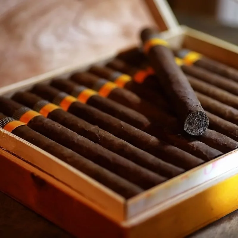 A wooden box containing multiple cigars, with one cigar partially removed and placed on top of the others.