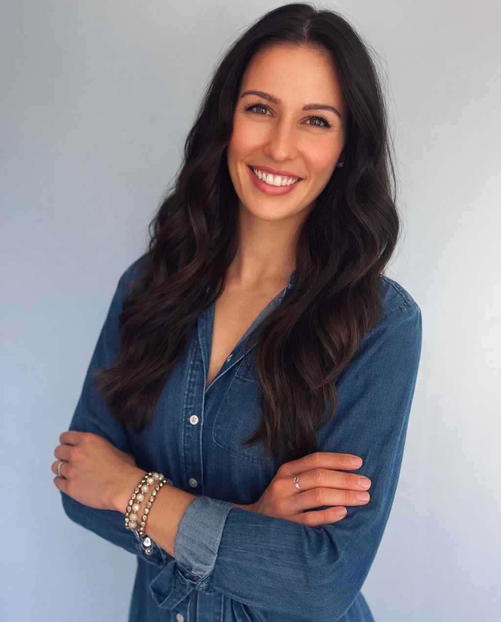A woman with long, wavy dark hair, smiling, wearing a denim shirt and jewelry, standing against a plain background.