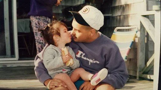 A young girl and a man sharing a kiss outdoors on a wooden deck.