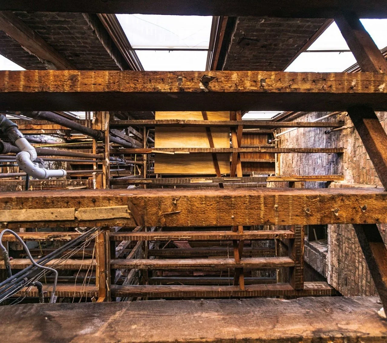 View of old wooden stairs with metal pipes and brick walls, looking upward from below.