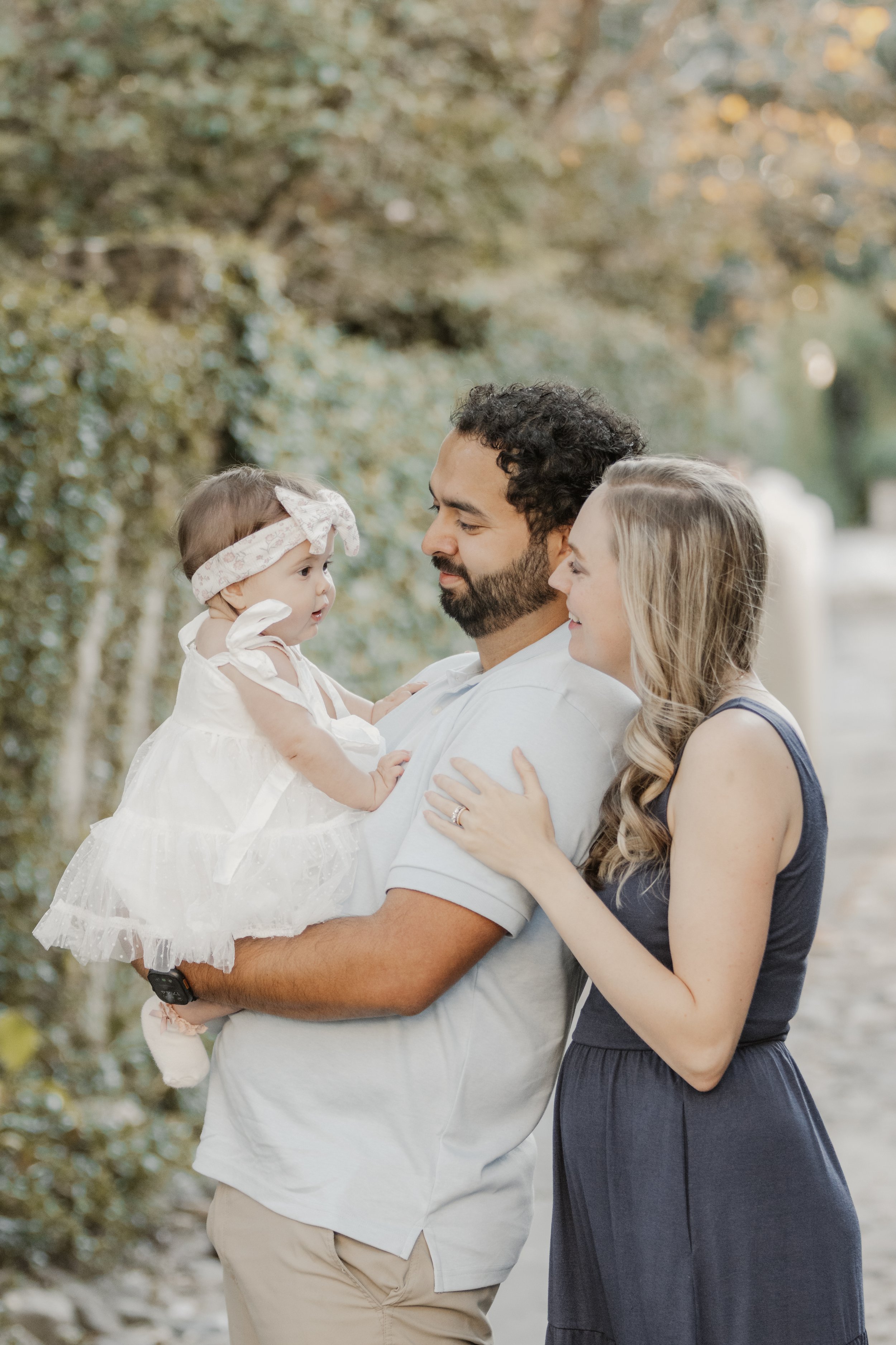 A family of three outdoors, with a man holding a young girl in a white dress and headband, and a woman standing beside them, all smiling and looking at each other, surrounded by trees with fall foliage.