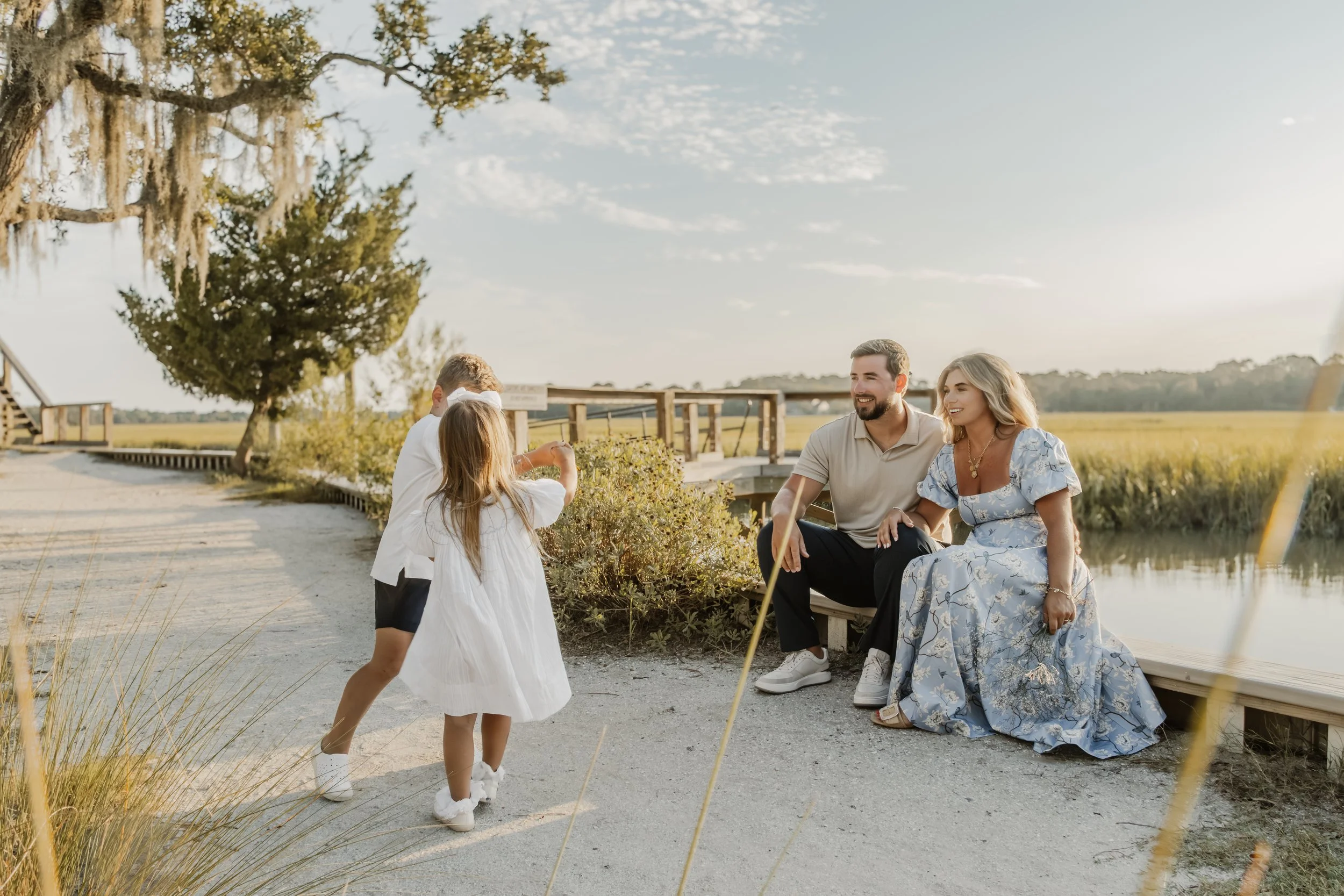A family of four enjoying along a paved trail near a waterway, with trees and a wooden bridge in the background during the daytime.