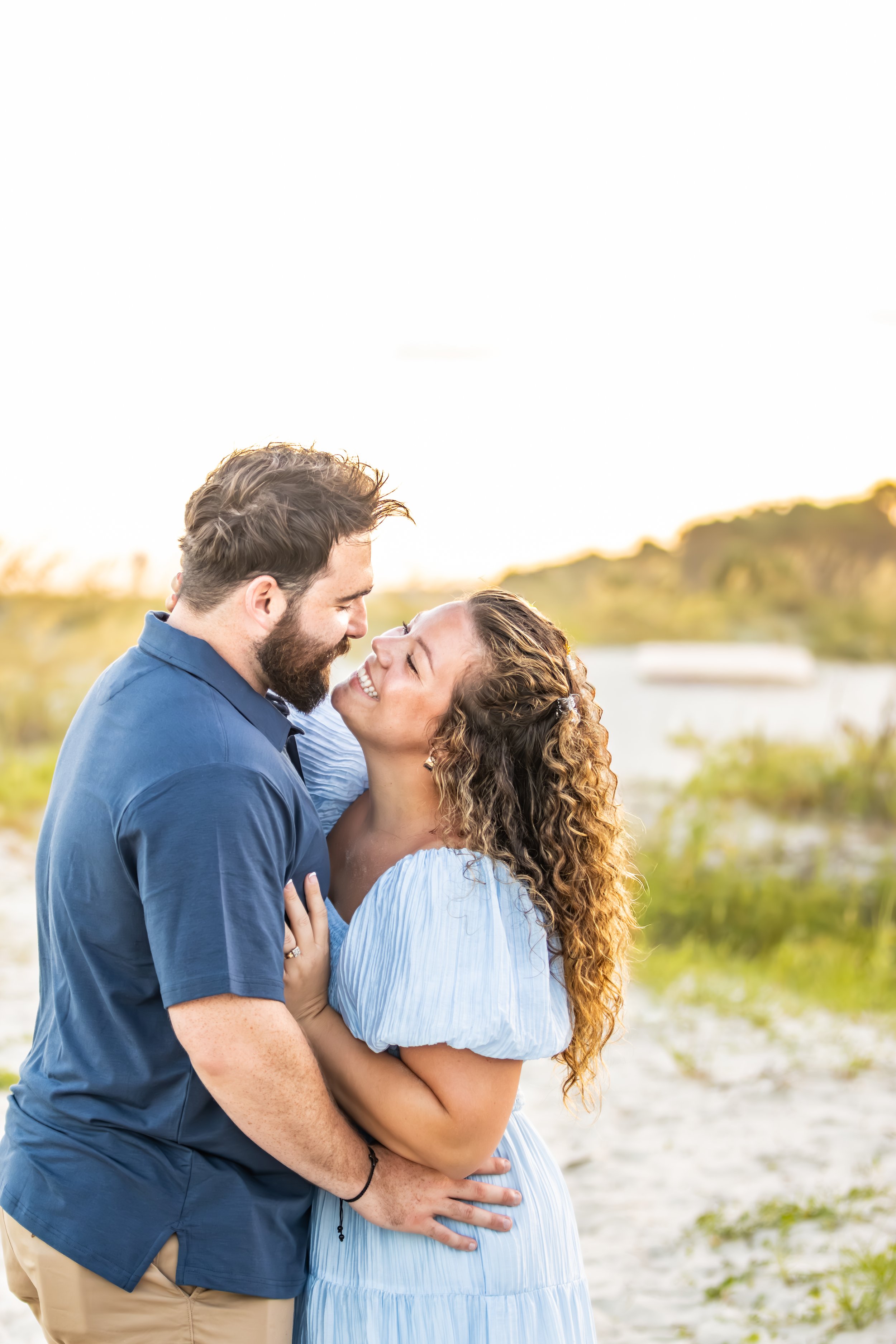 A couple embraces each other on a beach at sunset, smiling and looking into each other's eyes.