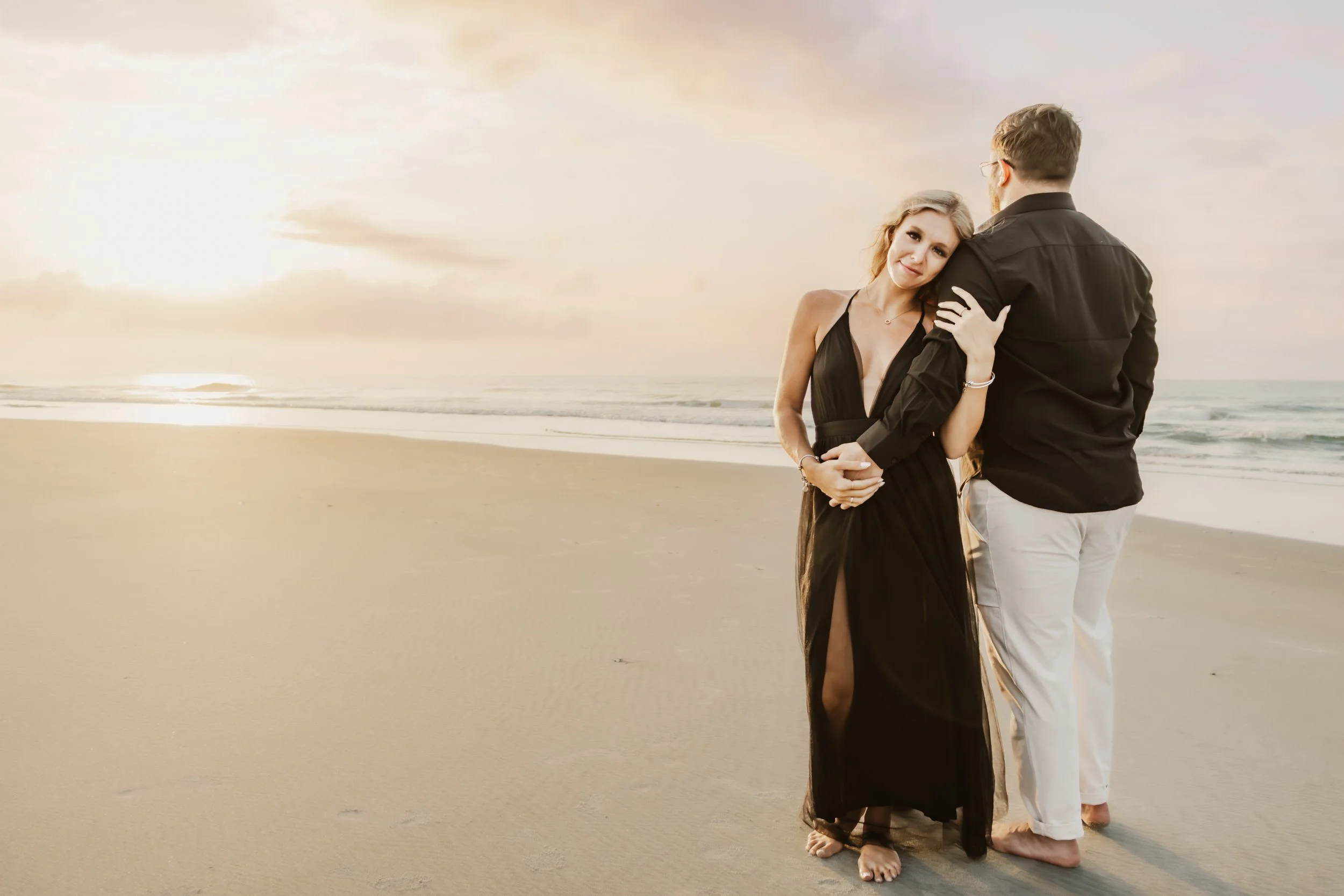 A couple standing on the beach at sunset, the woman is wearing a black dress and the man is in a black shirt and white pants, embracing each other.