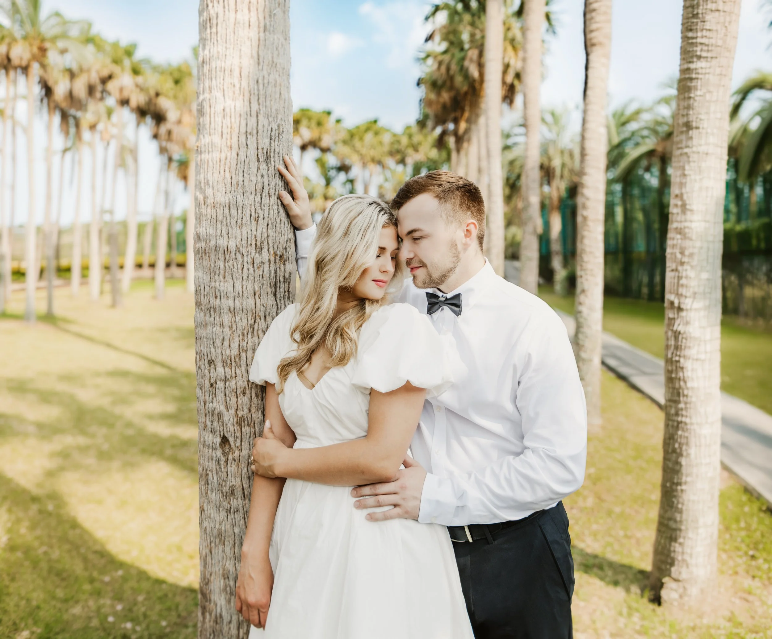A young couple in wedding attire sharing a tender moment outdoors among palm trees, with the woman resting her head against the man's forehead.