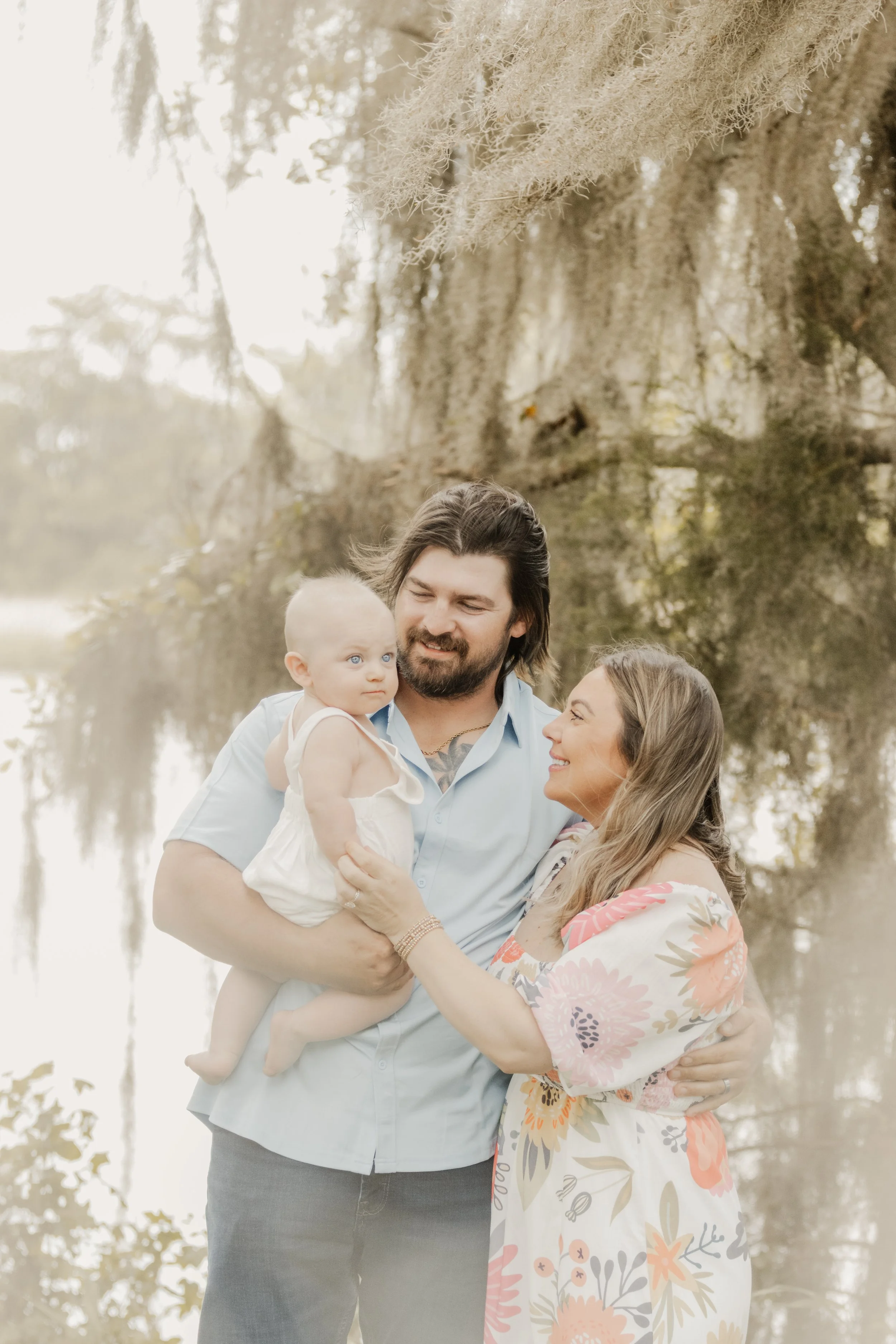 A family of three, including a man, a woman, and a baby, standing outdoors near a lake with hanging moss and trees in the background. The man is holding the baby, and the woman is smiling at them.