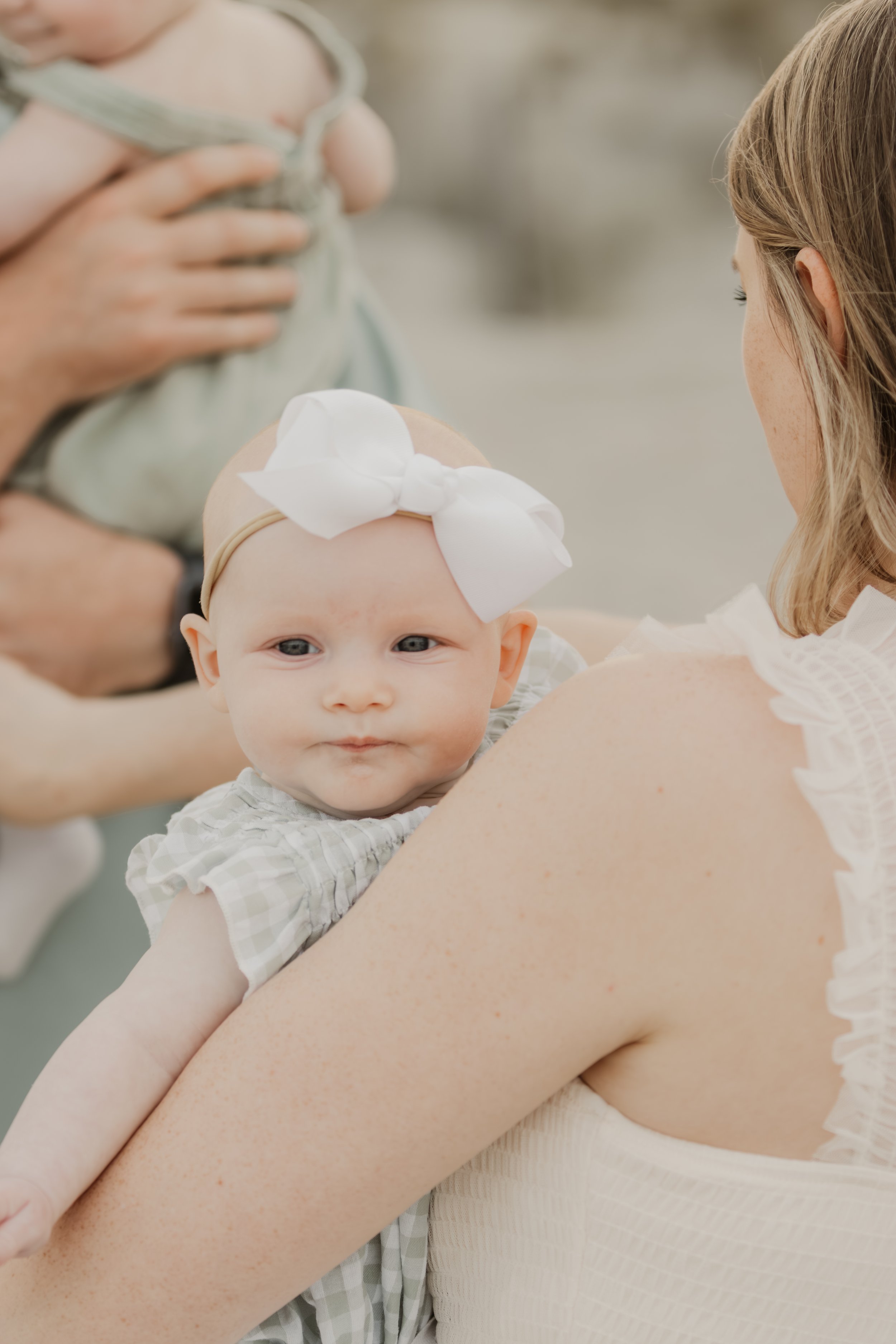 A baby girl with a white bow headband resting on a woman's shoulder during a special occasion, with a person holding another baby in the background.