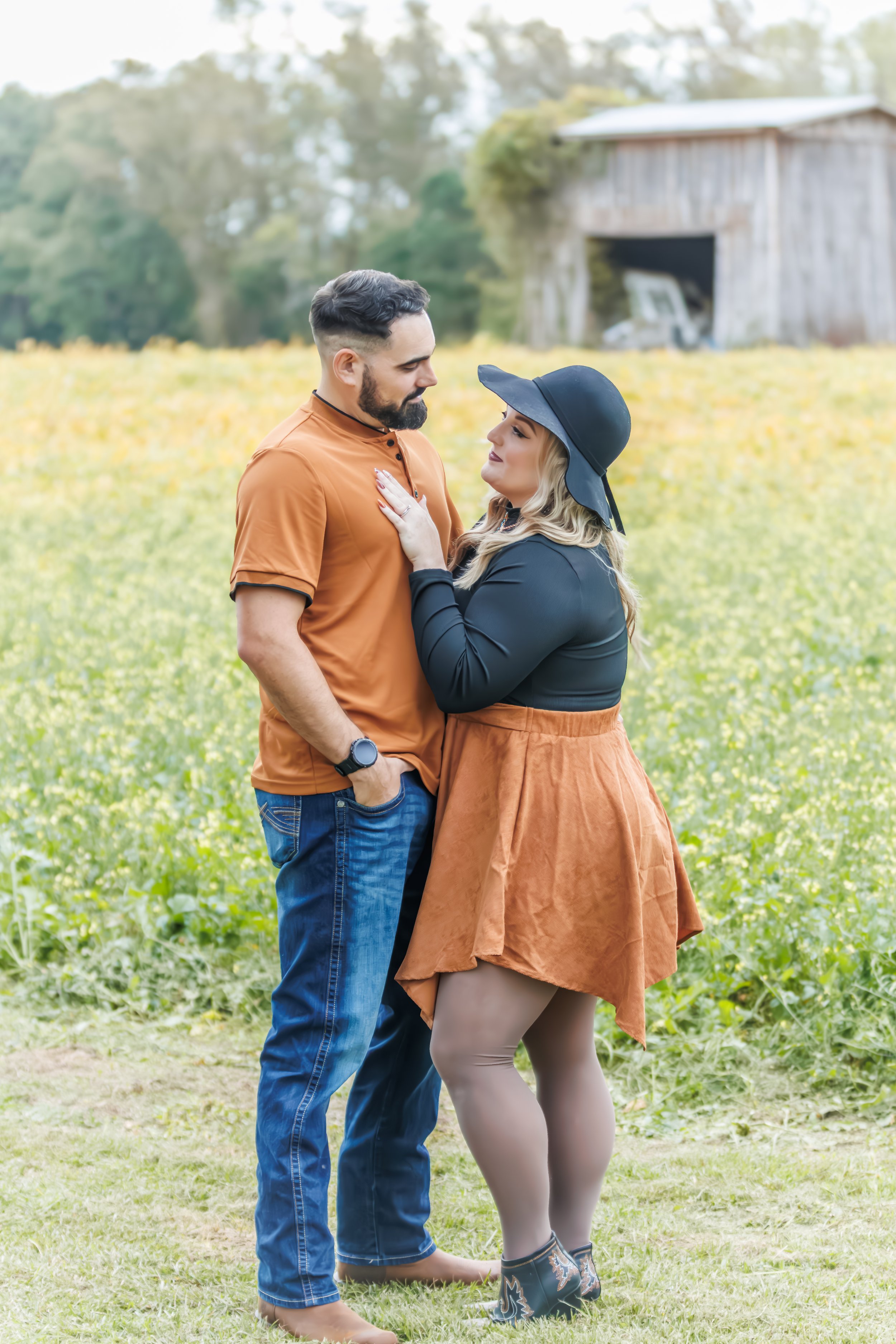 A couple standing close together outdoors in a field with an old barn in the background. The man has short dark hair and a beard, wearing a brown shirt and jeans, with his left hand in his pocket. The woman has long blond hair, wearing a black wide-b