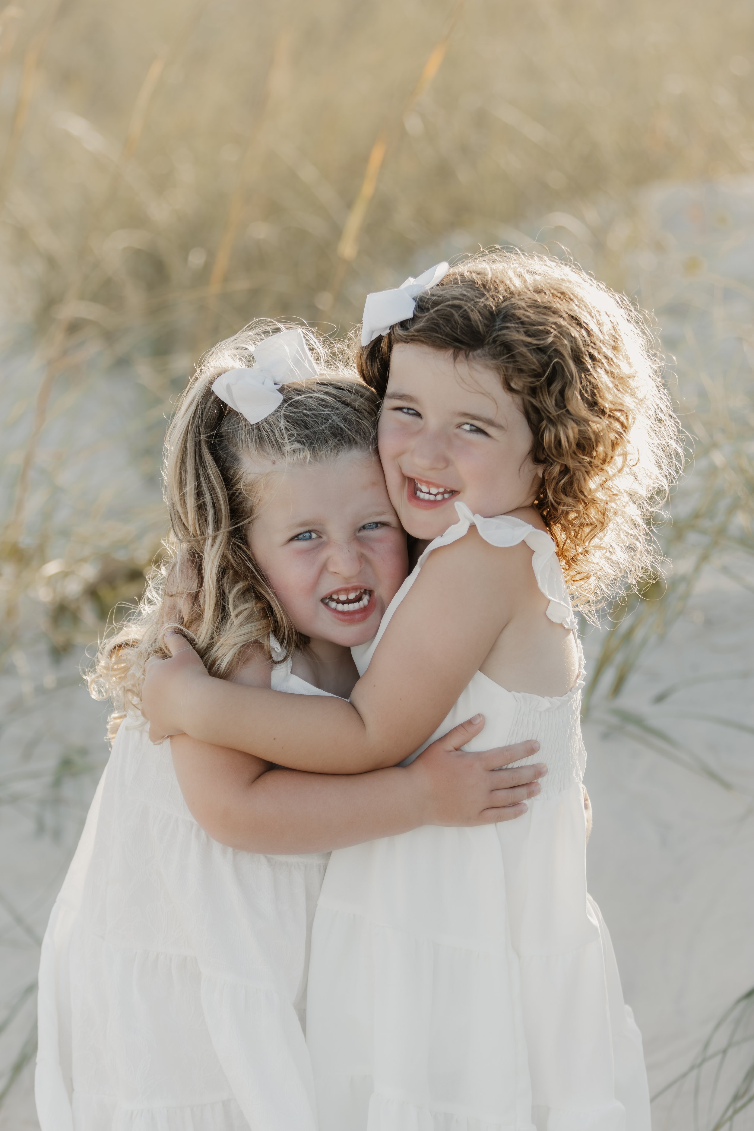 Two young girls with curly hair and white dresses hugging each other on a beach, background of dry reeds.