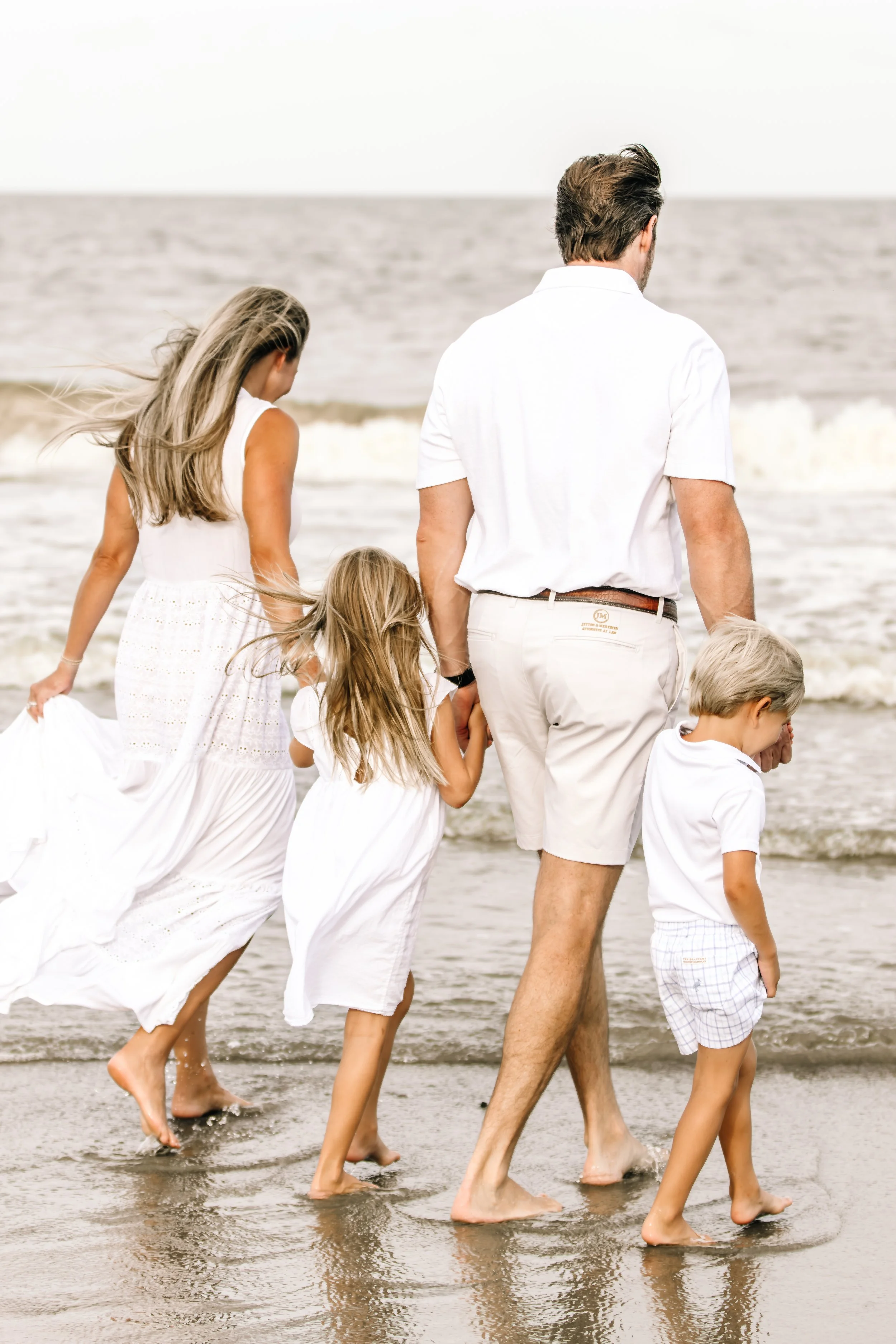 Family walking along the beach near the shoreline, dressed in white summer clothes, with the ocean in the background.