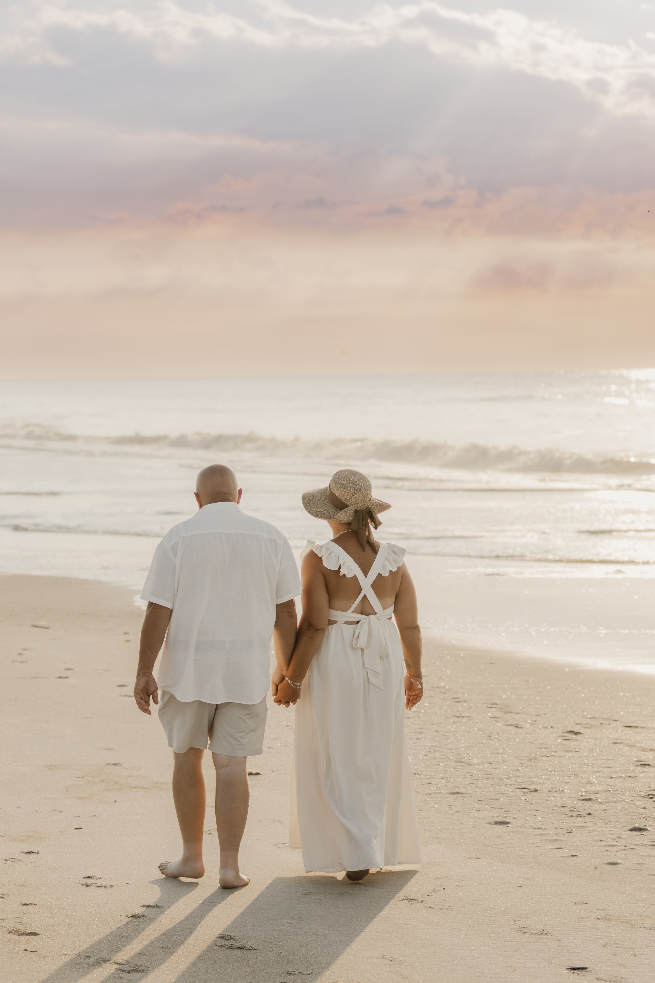 A couple walking hand in hand on the beach at sunset, dressed in white clothes. The woman is wearing a large sun hat and a long dress, and the man is in a short-sleeved shirt and shorts.
