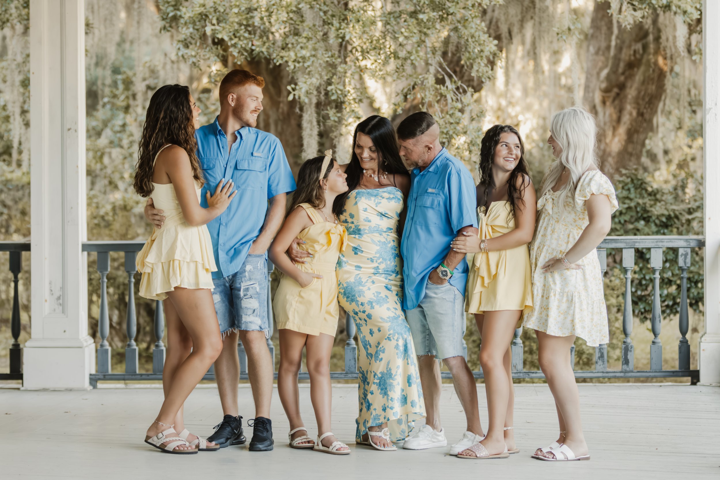 Family group on porch in summer, including three women, three men, and two young girls, all smiling and dressed in casual, light-colored clothing.