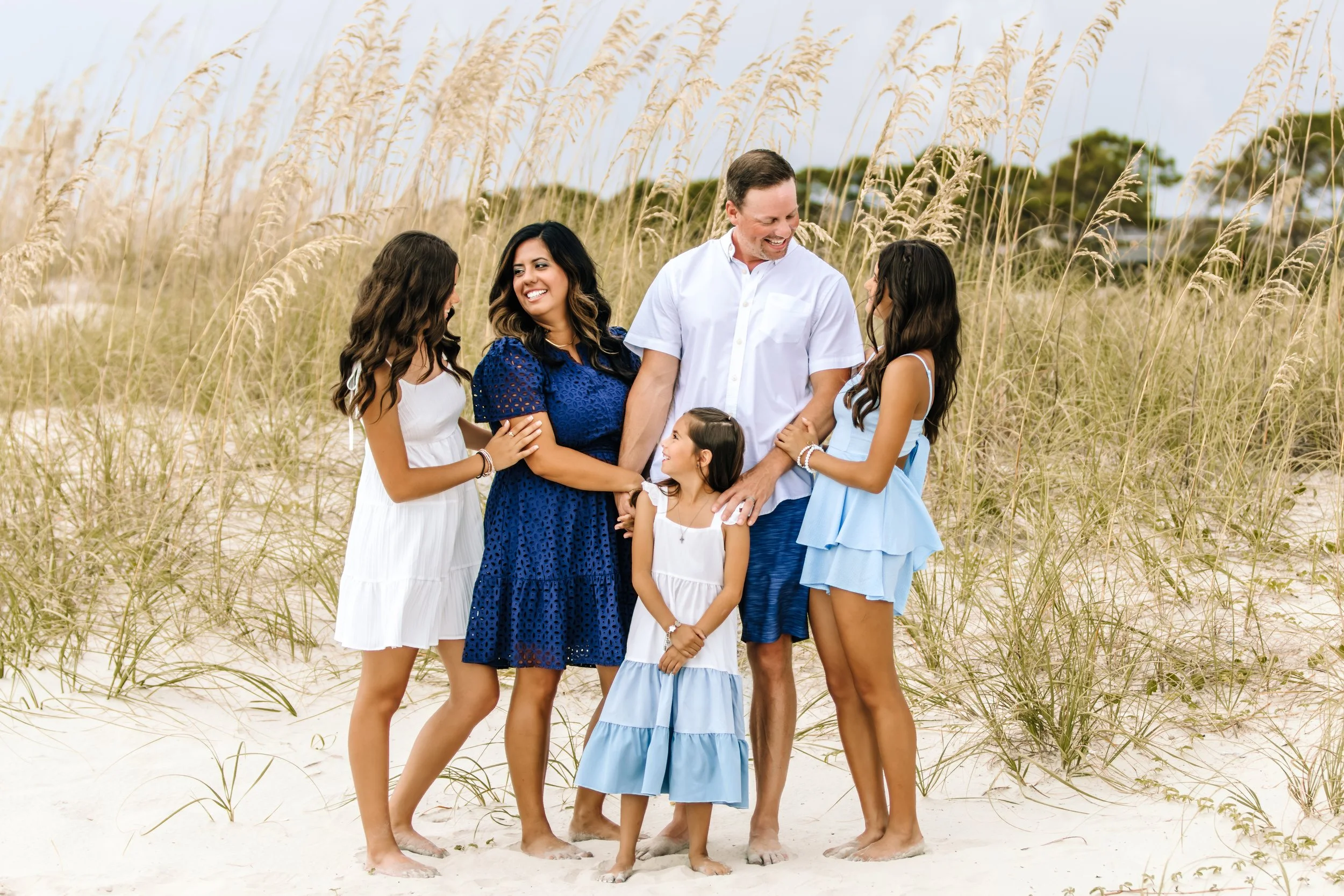 A family of six standing on a sandy beach with tall grass, smiling and holding hands, dressed in white and blue summer outfits.