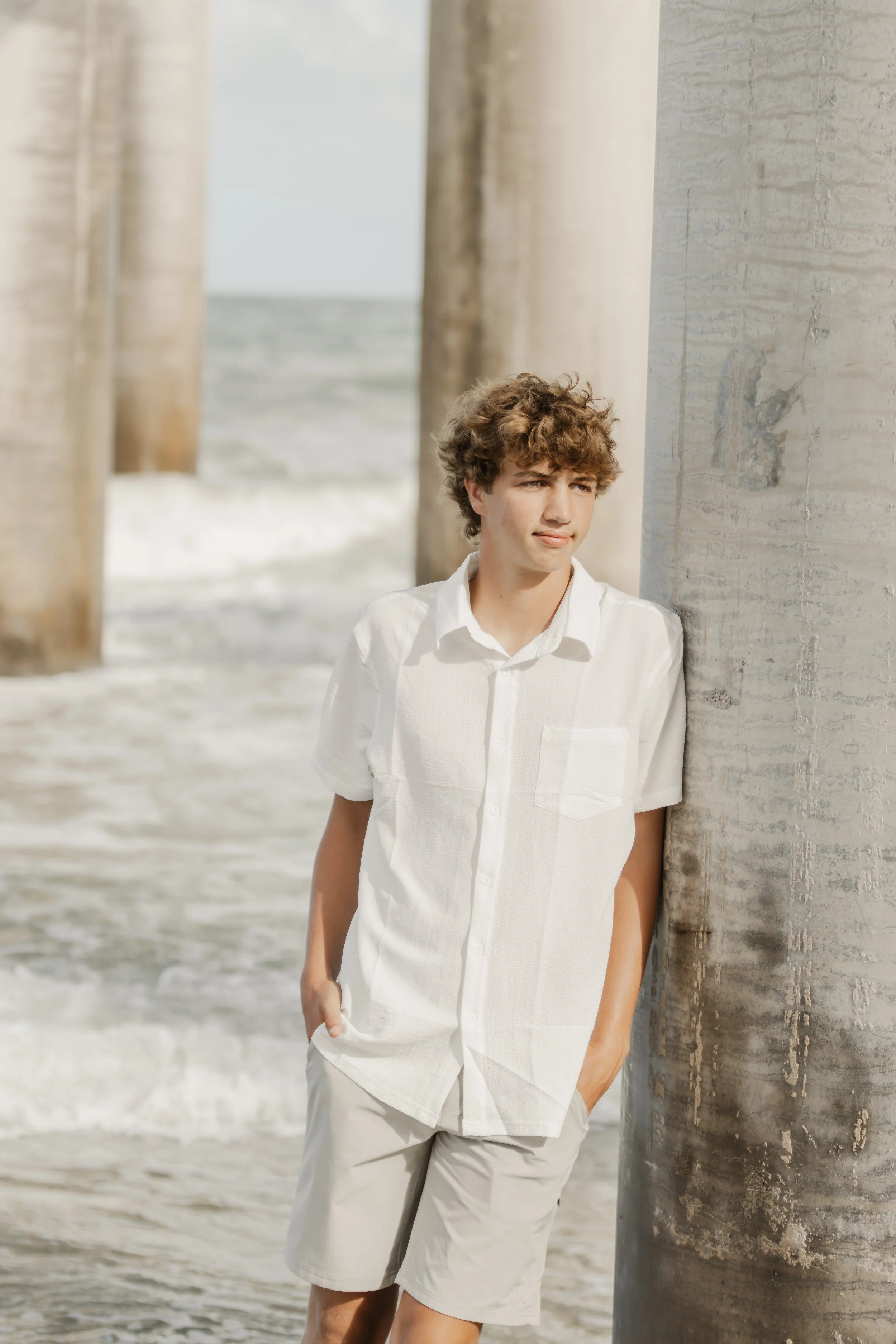 A young man with curly hair wearing a white shirt and beige shorts leaning against a concrete pillar at the beach with ocean waves and a pier in the background.