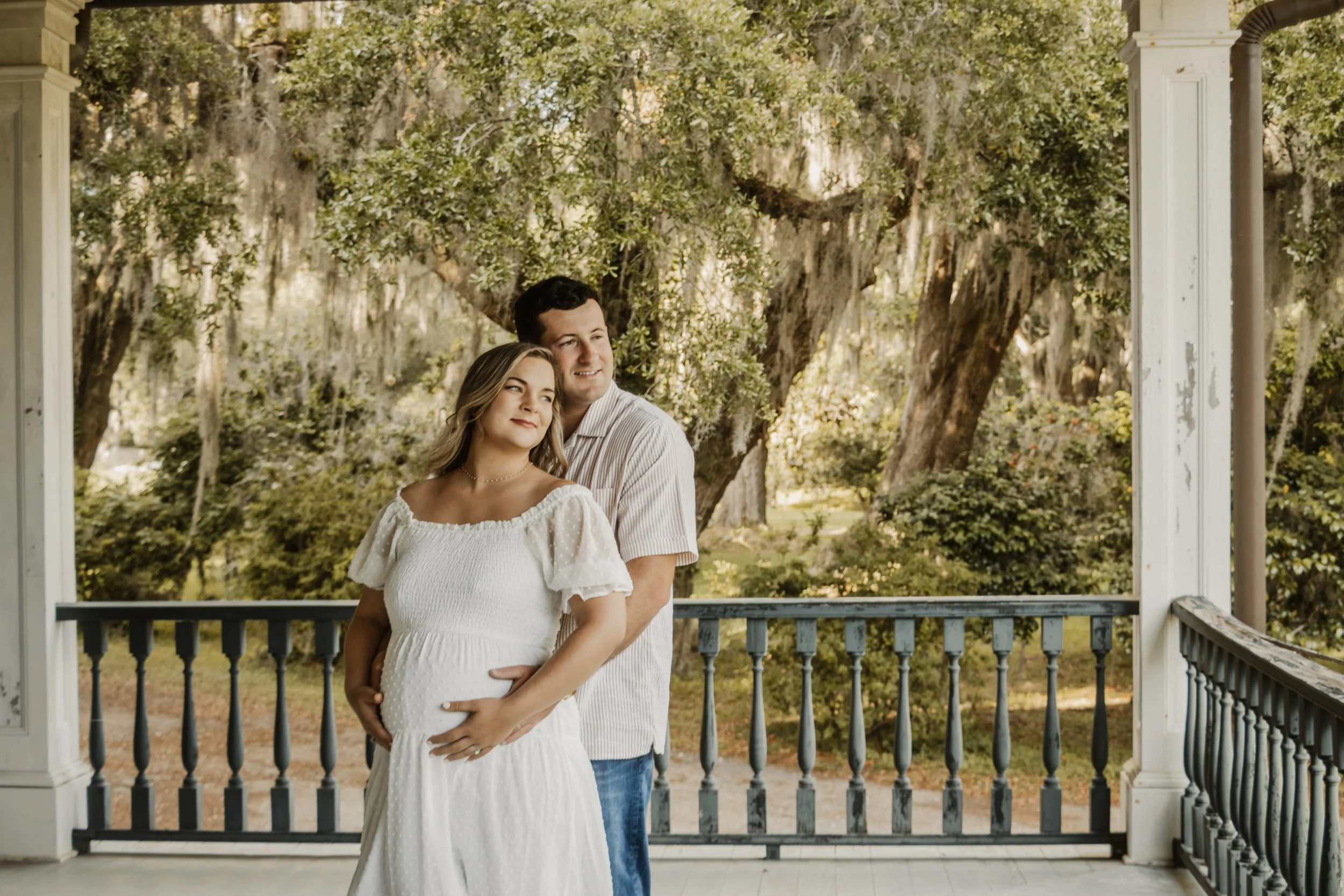 A pregnant woman and a man standing on a porch with trees in the background. The woman is wearing a white dress, and the man is in a striped shirt and jeans. They are looking outward, with the man embracing the woman from behind.