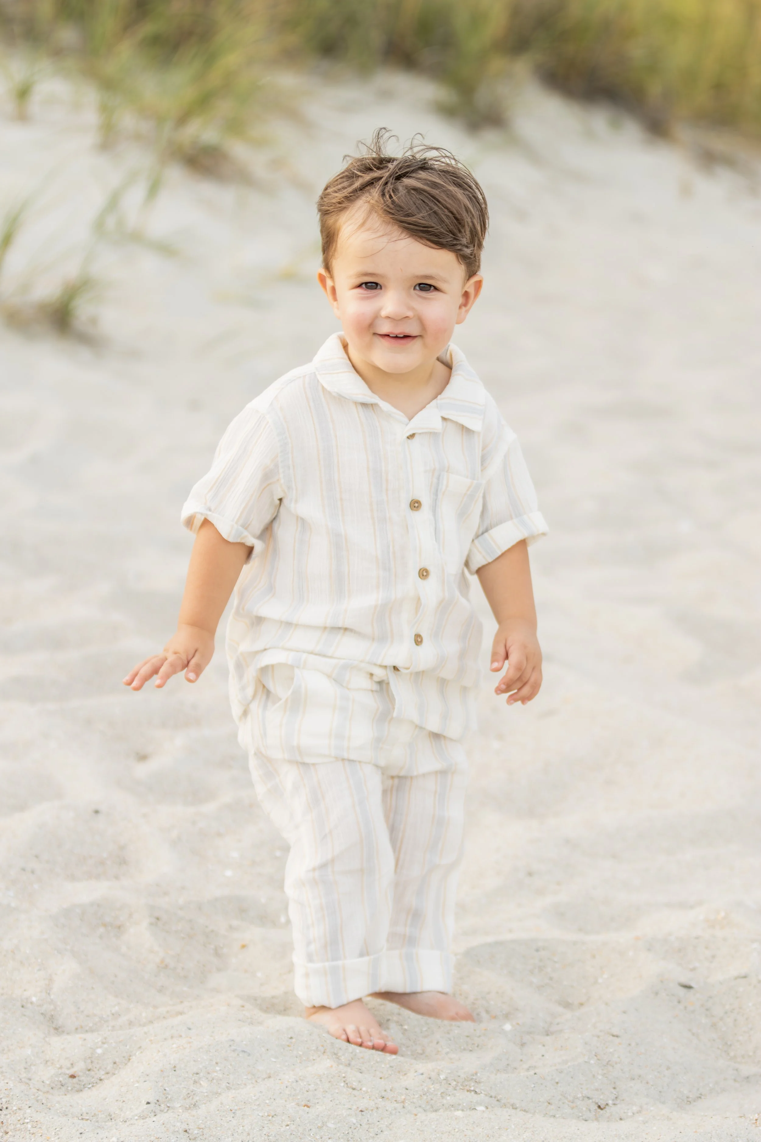 A young boy with brown hair and wearing a cream-colored striped outfit is walking barefoot on a sandy beach, smiling at the camera.
