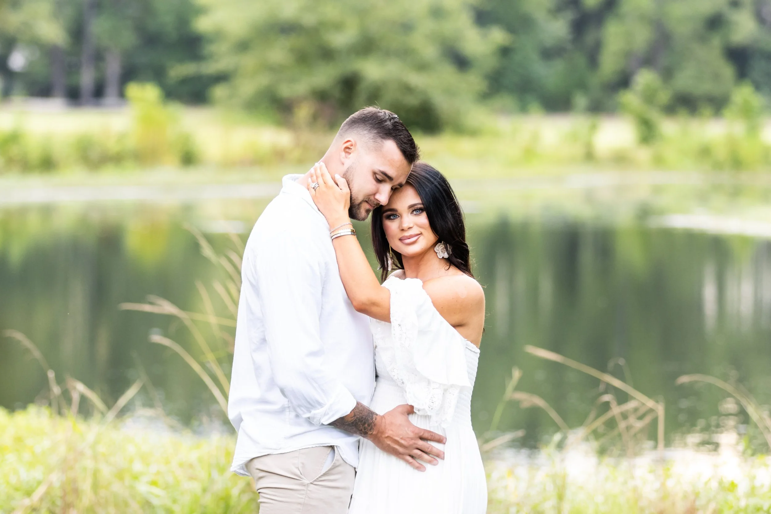 A loving couple standing by a lake, embracing each other with trees in the background.