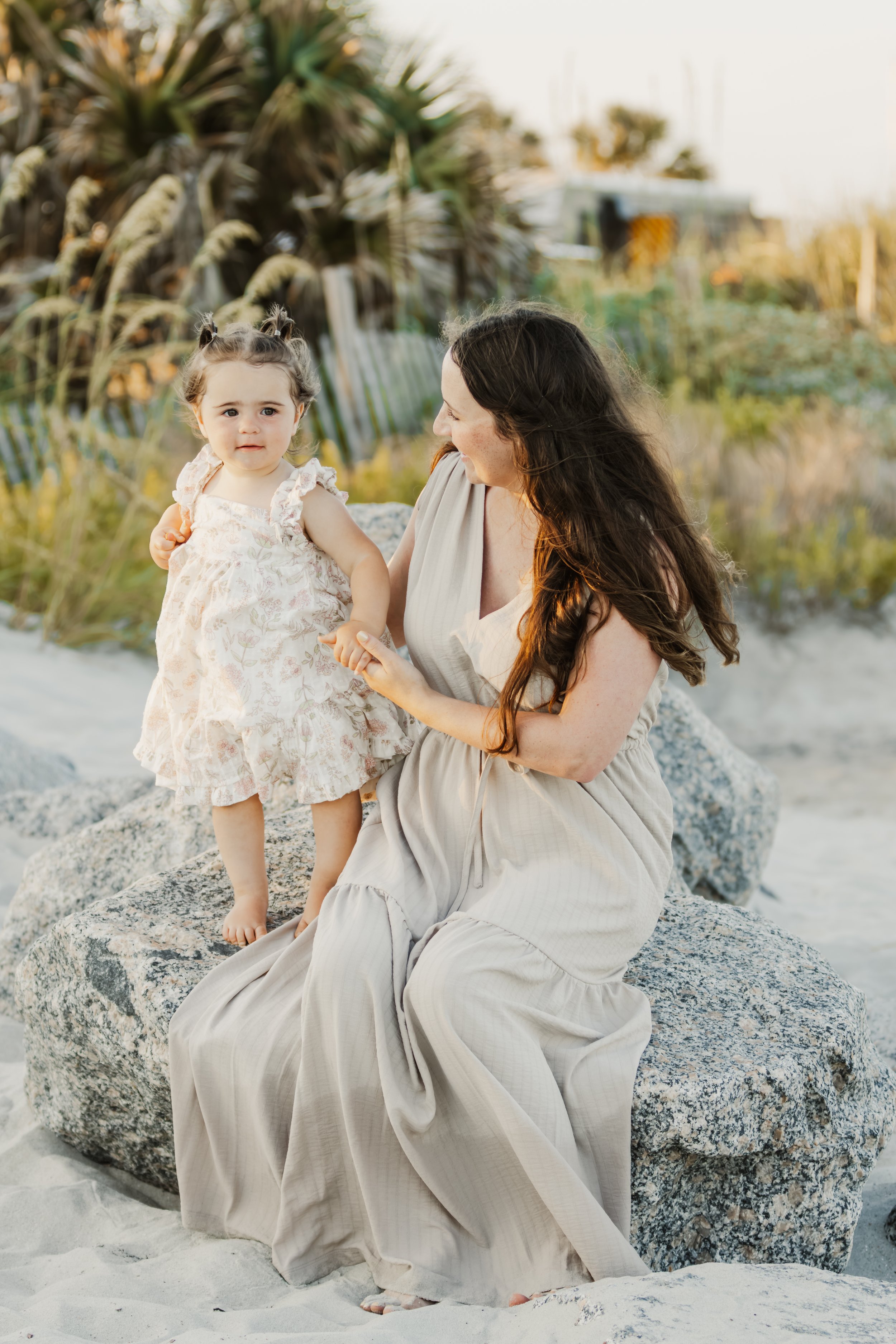 A woman and a young girl sitting on a large rock at the beach with sand, plants, and greenery in the background during sunset.