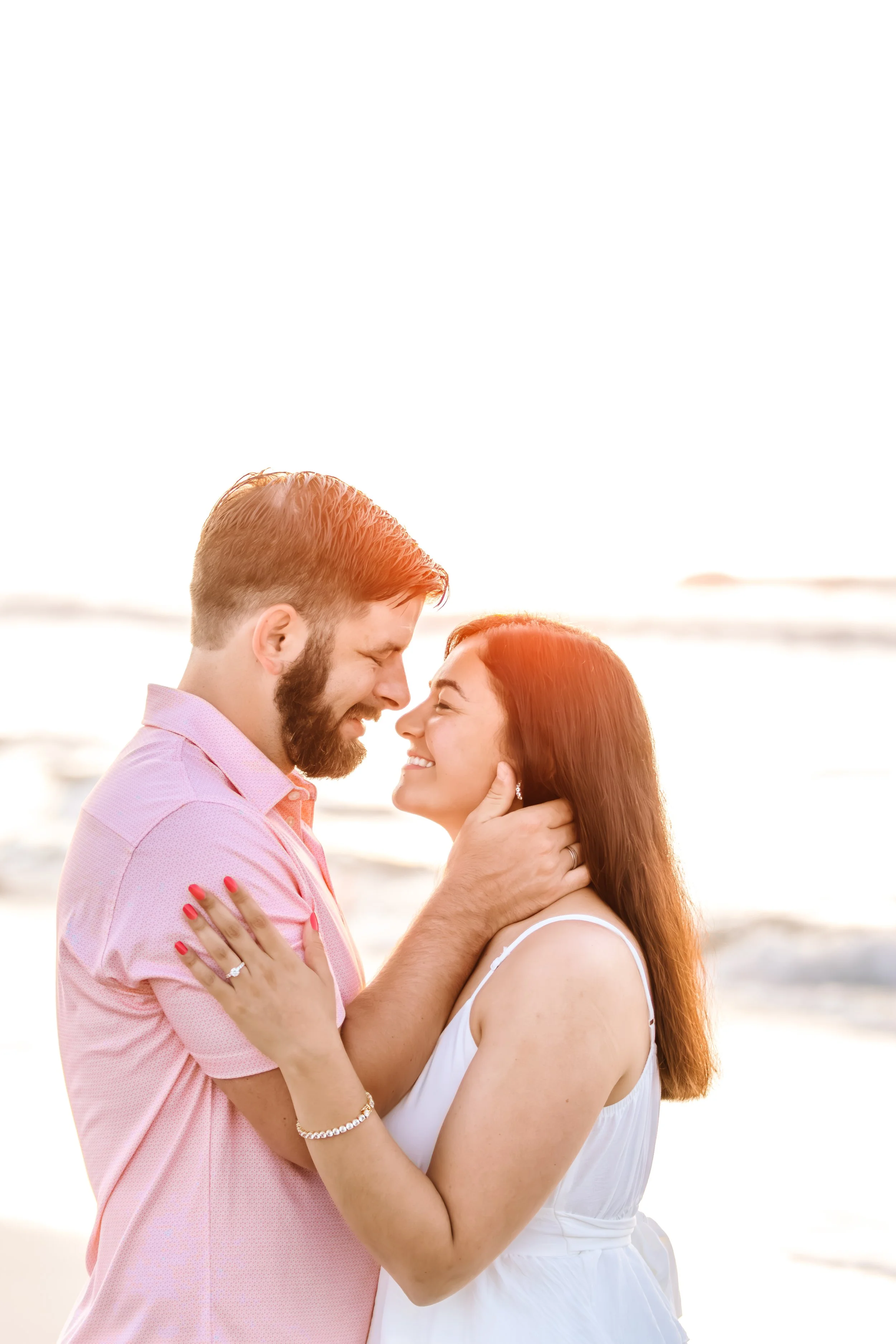 A couple smiling and embracing on the beach during sunset, with ocean waves in the background.