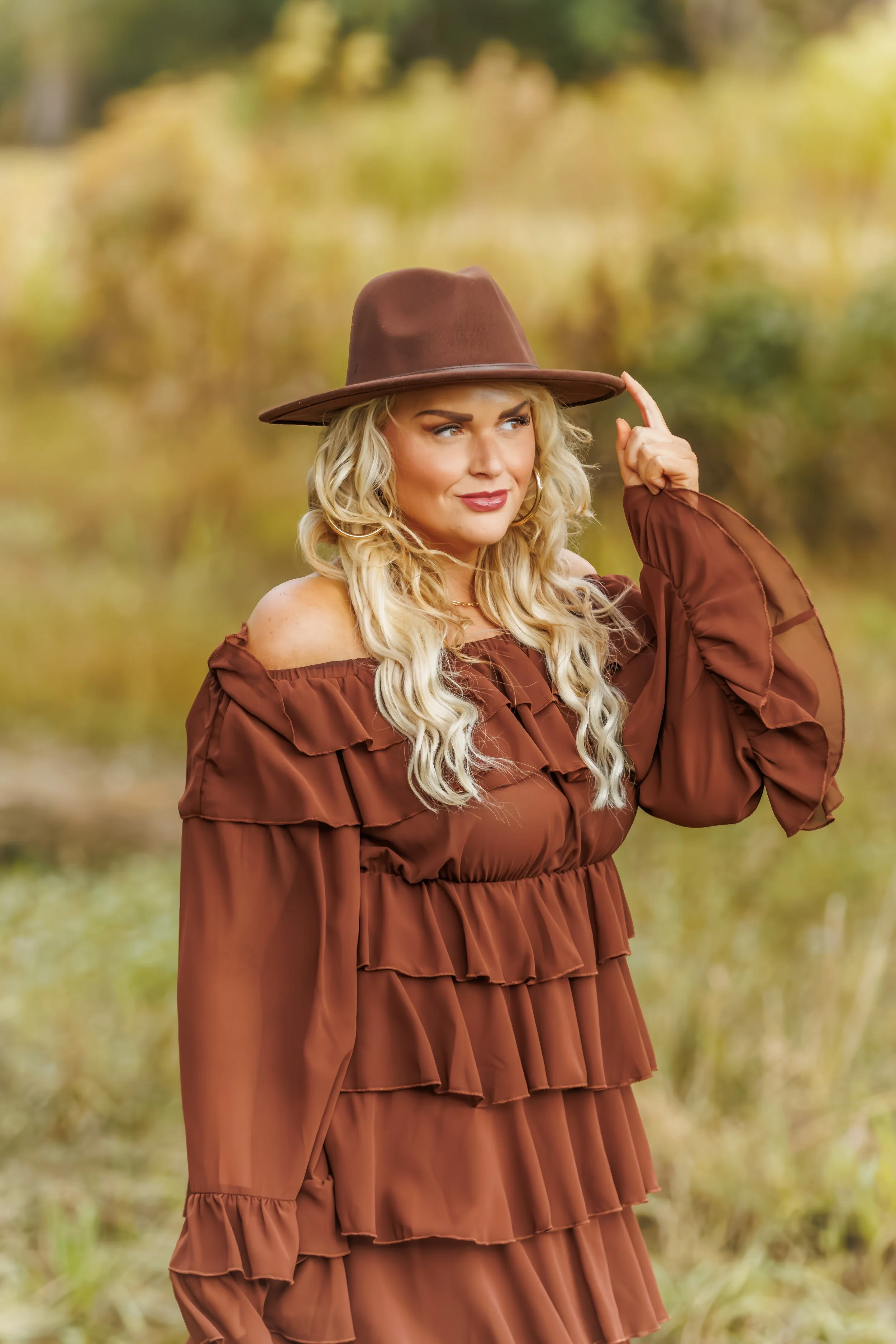 A woman with long, wavy blonde hair wearing a brown off-shoulder ruffled dress and a matching wide-brimmed hat, standing outdoors with blurred autumn trees in the background, touching her hat.