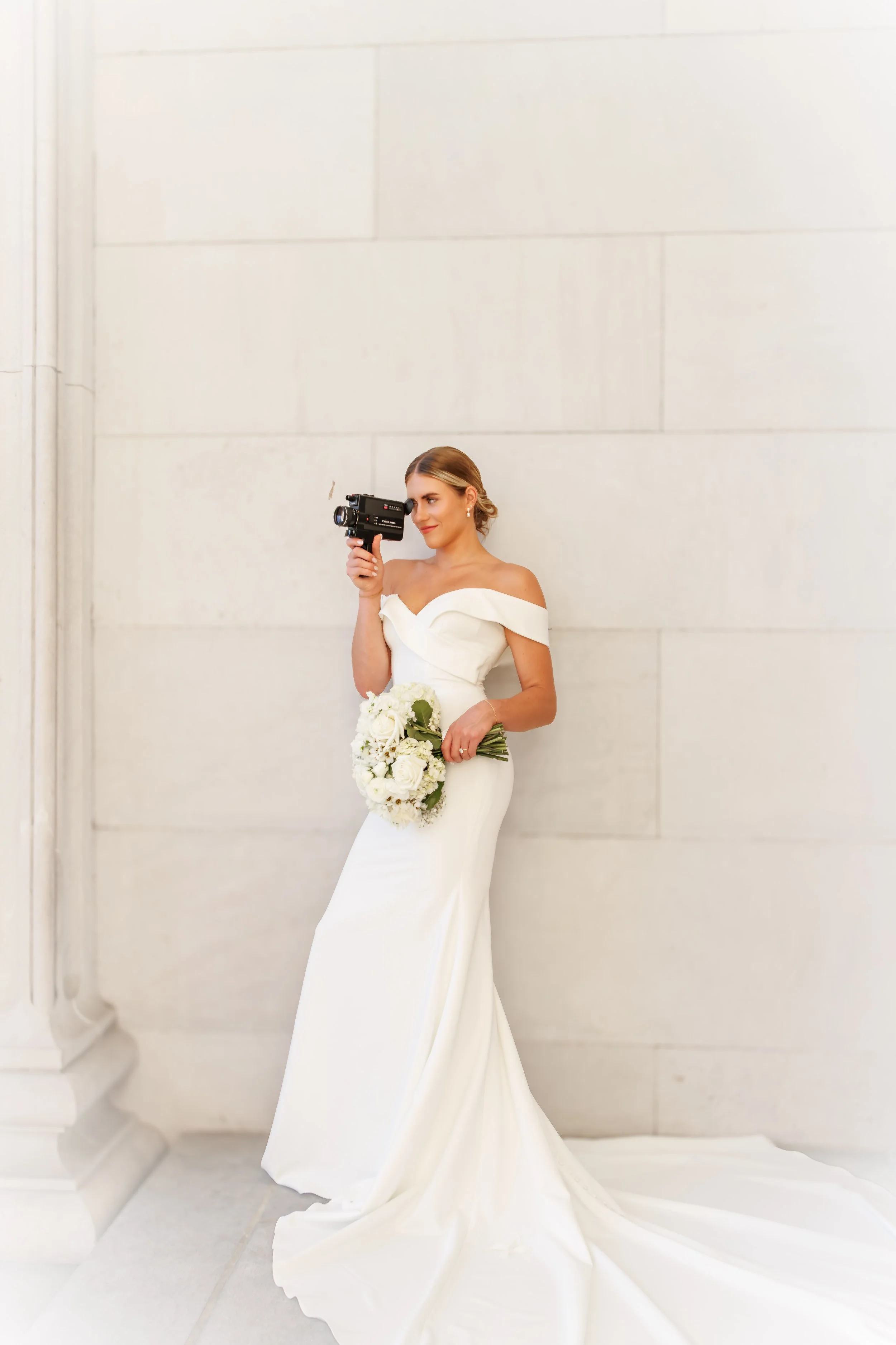 A bride in a white wedding gown holding a bouquet of white flowers, standing against a beige stone wall while looking through a vintage film camera.