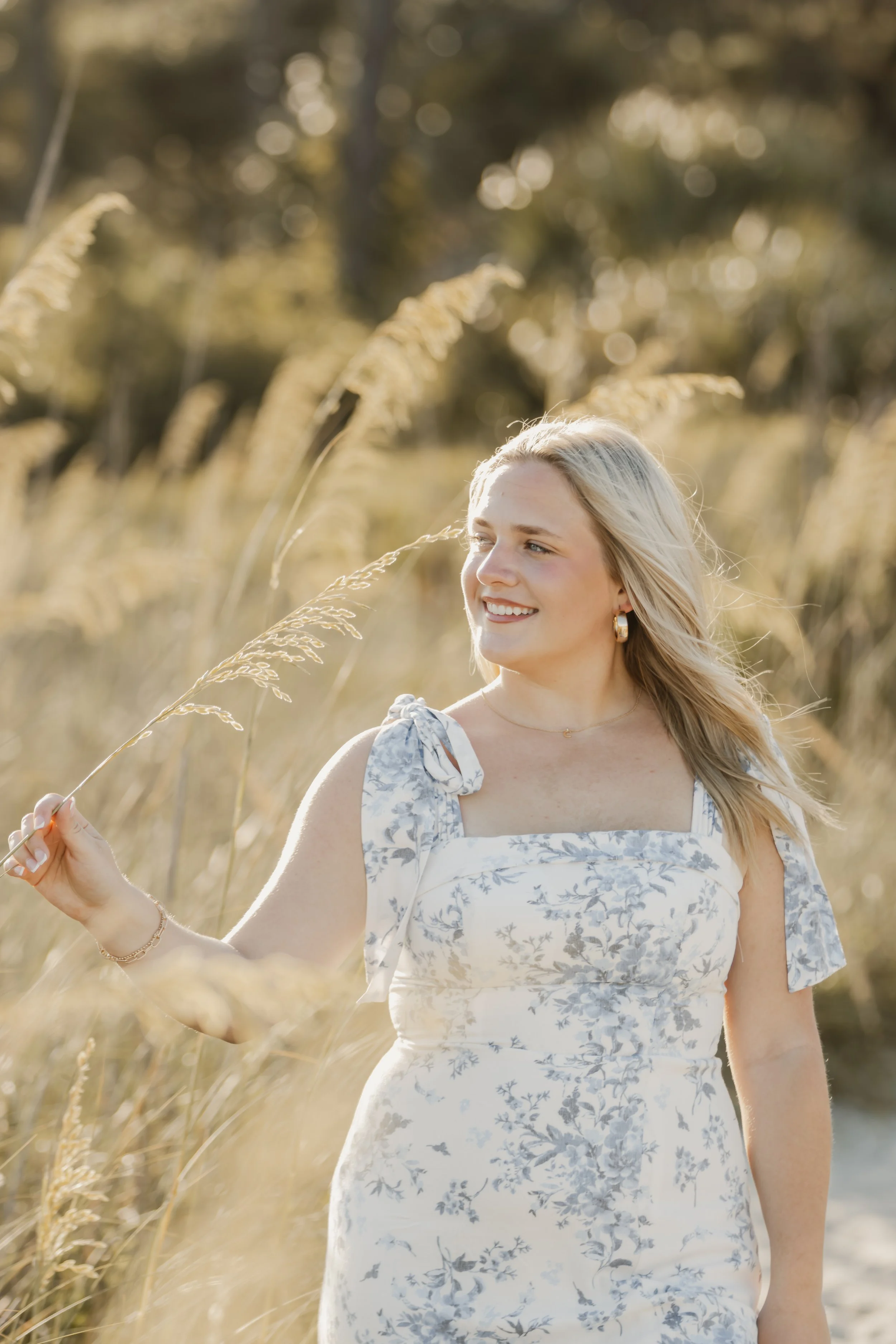 A young woman with blonde hair in a floral dress standing in a field of tall grass, illuminated by warm sunlight.