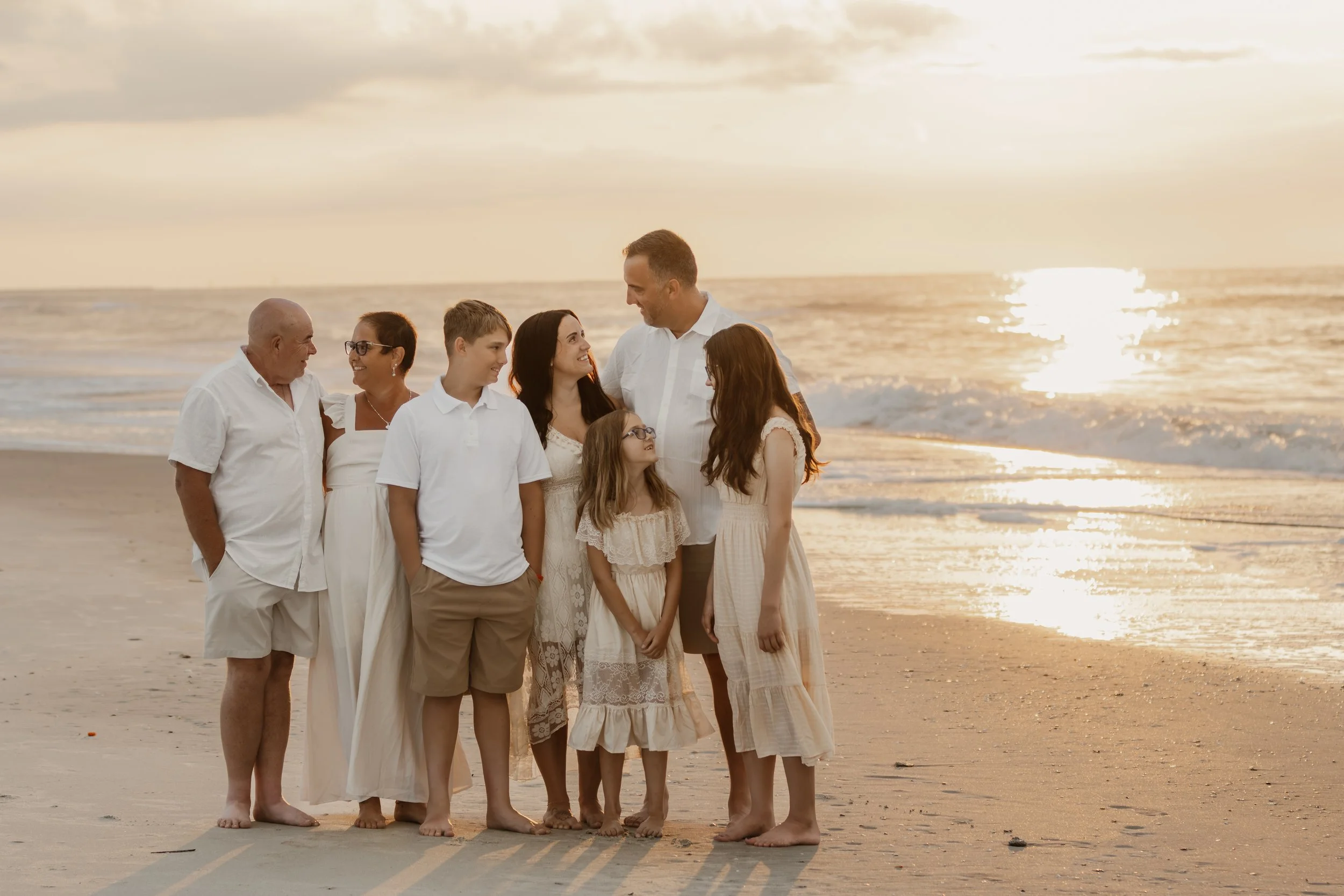 Family of nine members standing on a beach during sunset, wearing white or light-colored clothing, smiling and looking at each other.