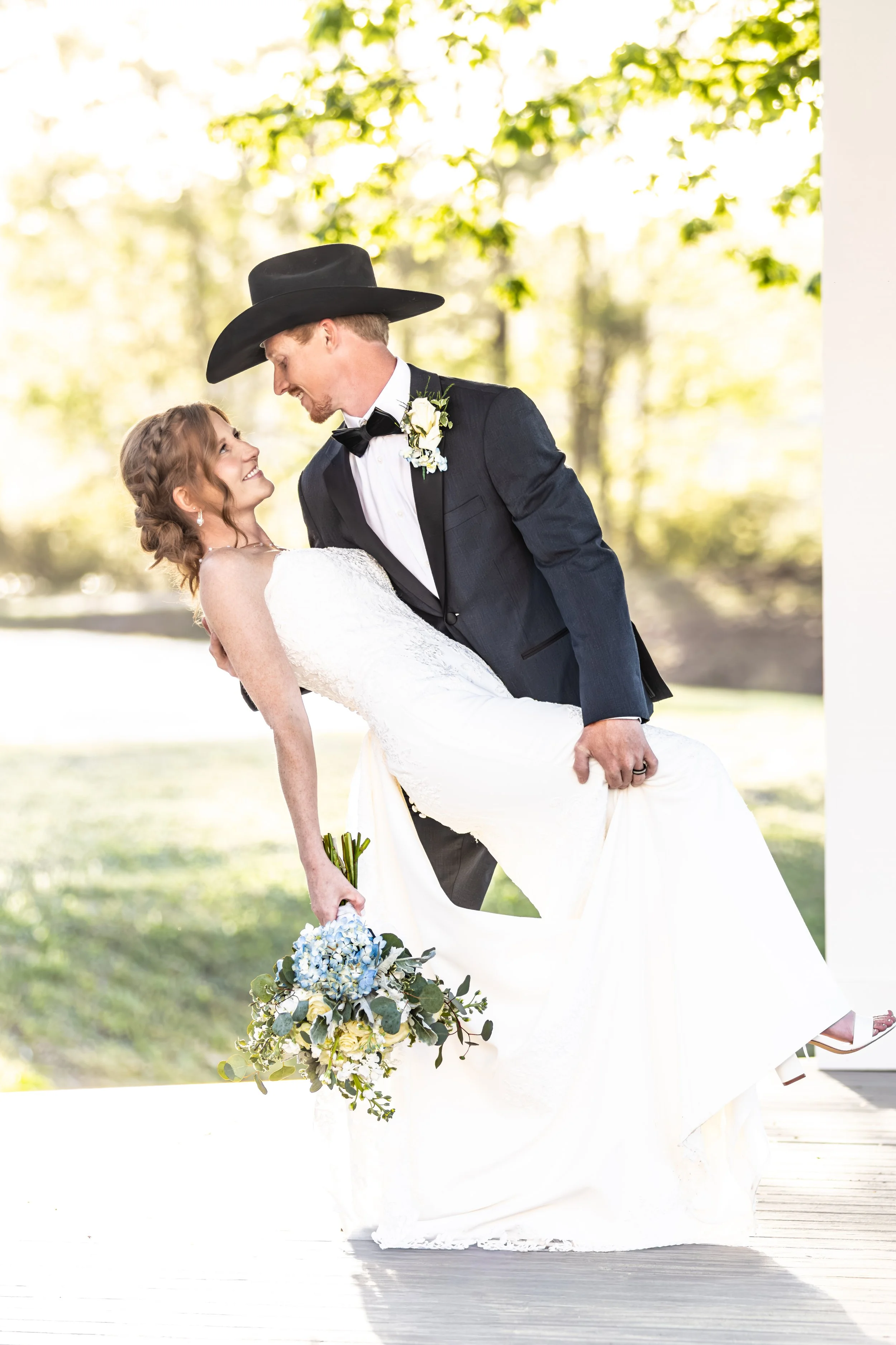 A groom in a black suit, bow tie, and cowboy hat holding a bride in a white wedding dress, outdoors with green trees in the background, during a wedding celebration.