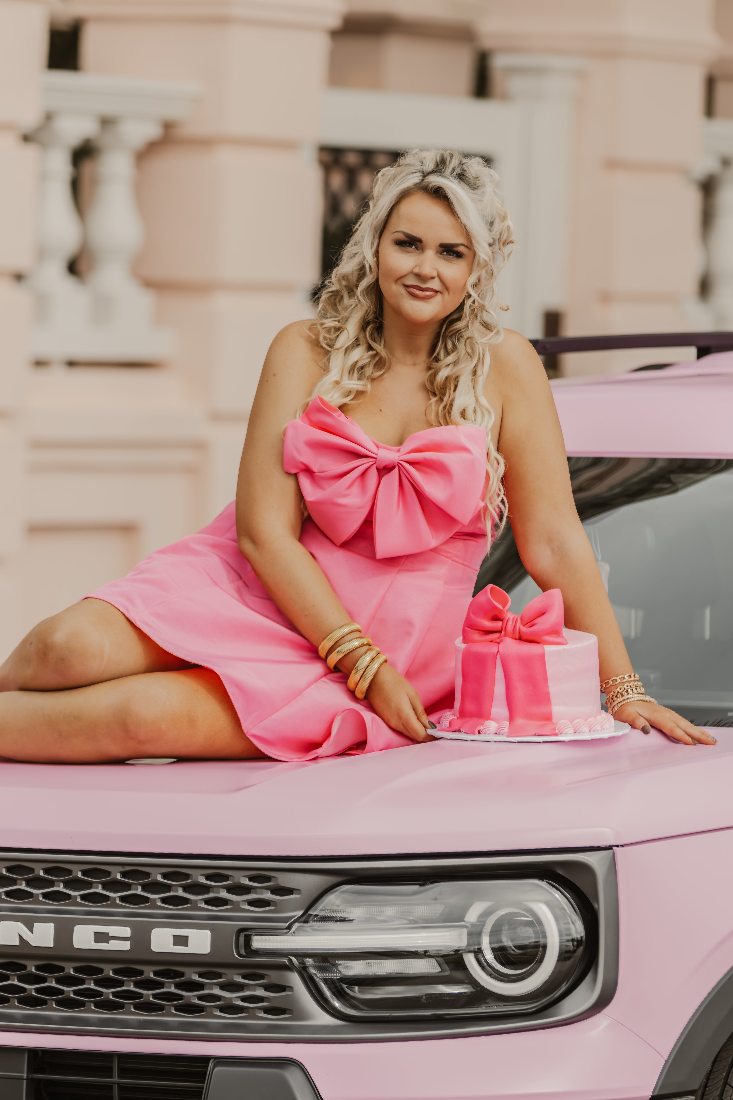 Woman in a pink dress with a large bow detail, sitting on the hood of a pink vehicle next to a pink birthday cake with a bow decoration.