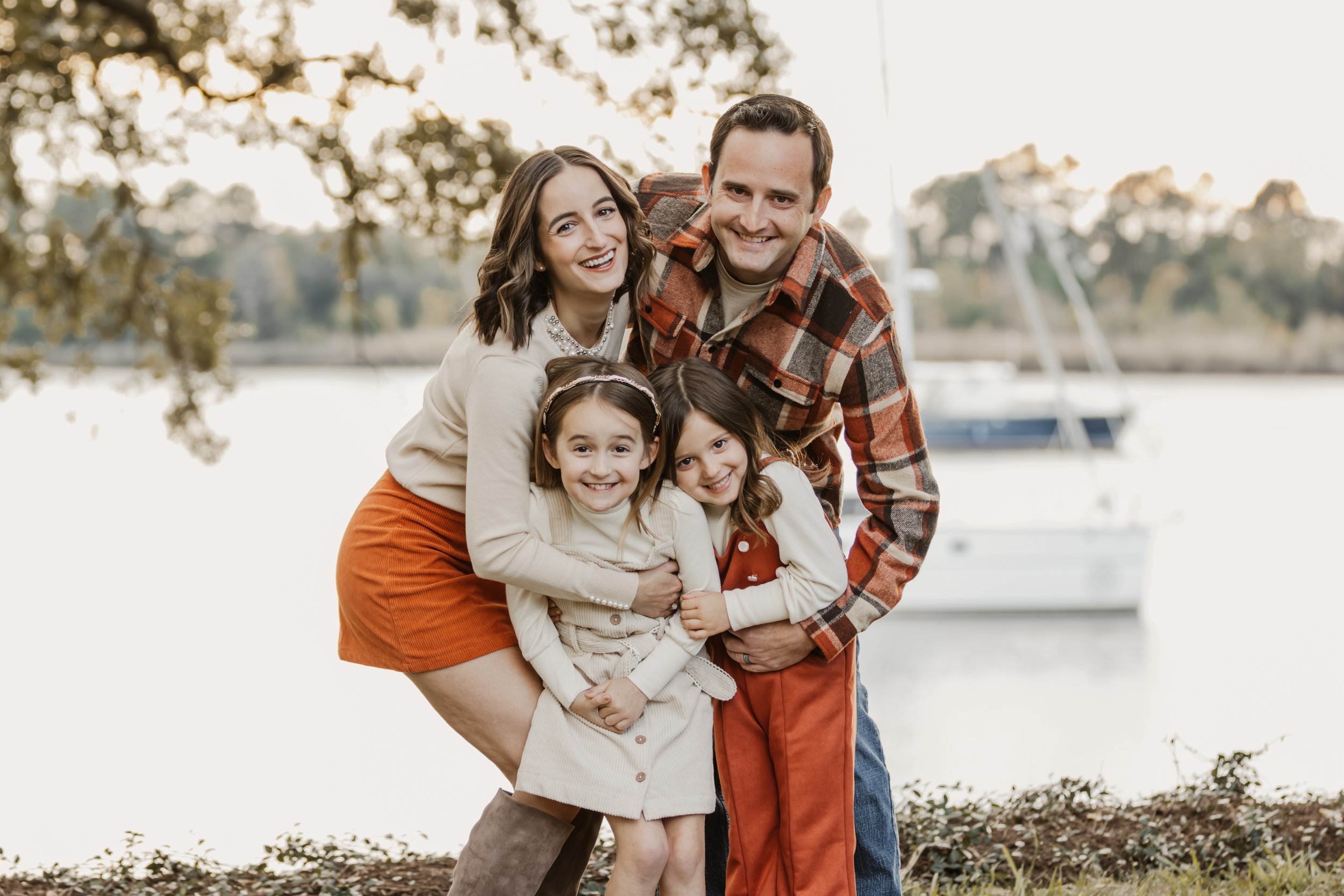 A happy family of four outdoors near a lake with a sailboat in the background, smiling and hugging.