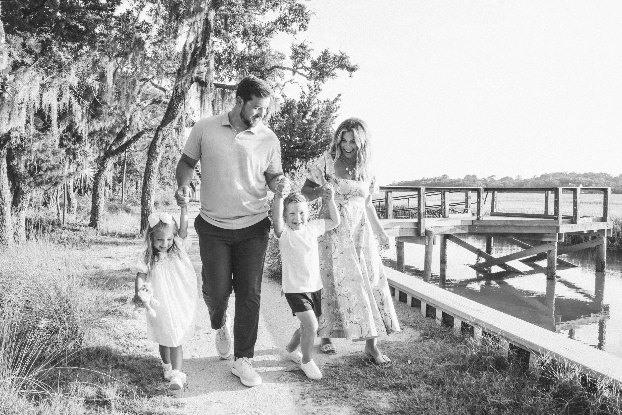 A family of four walking along a trail near a body of water, holding hands and smiling. The group includes a man, a woman, and two young girls, with trees and a dock visible in the background.