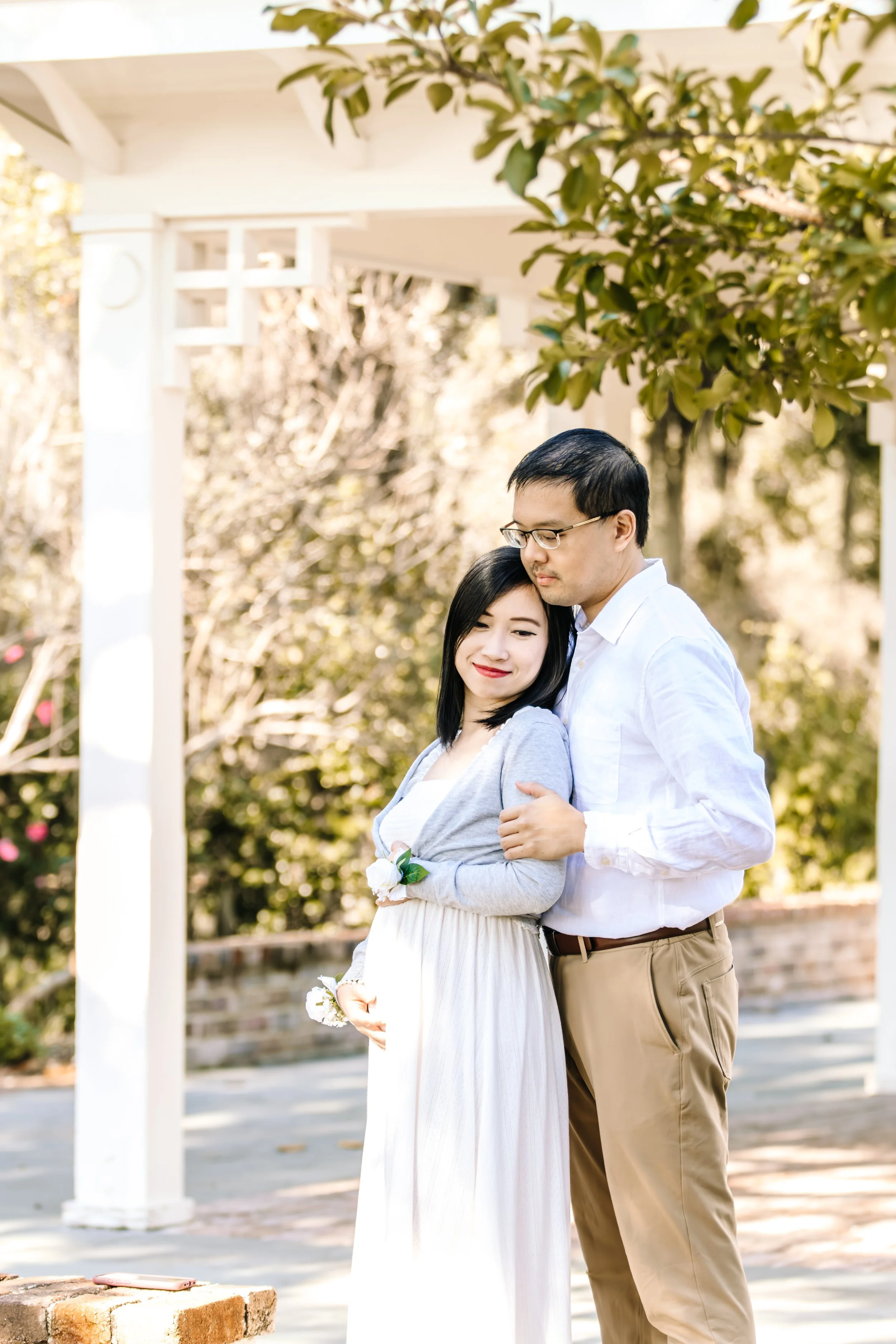 A pregnant woman and a man stand close together outdoors, with the woman smiling and the man gently holding her. They are under a white pergola, with trees and bushes in the background, suggesting a garden or park setting on a sunny day.
