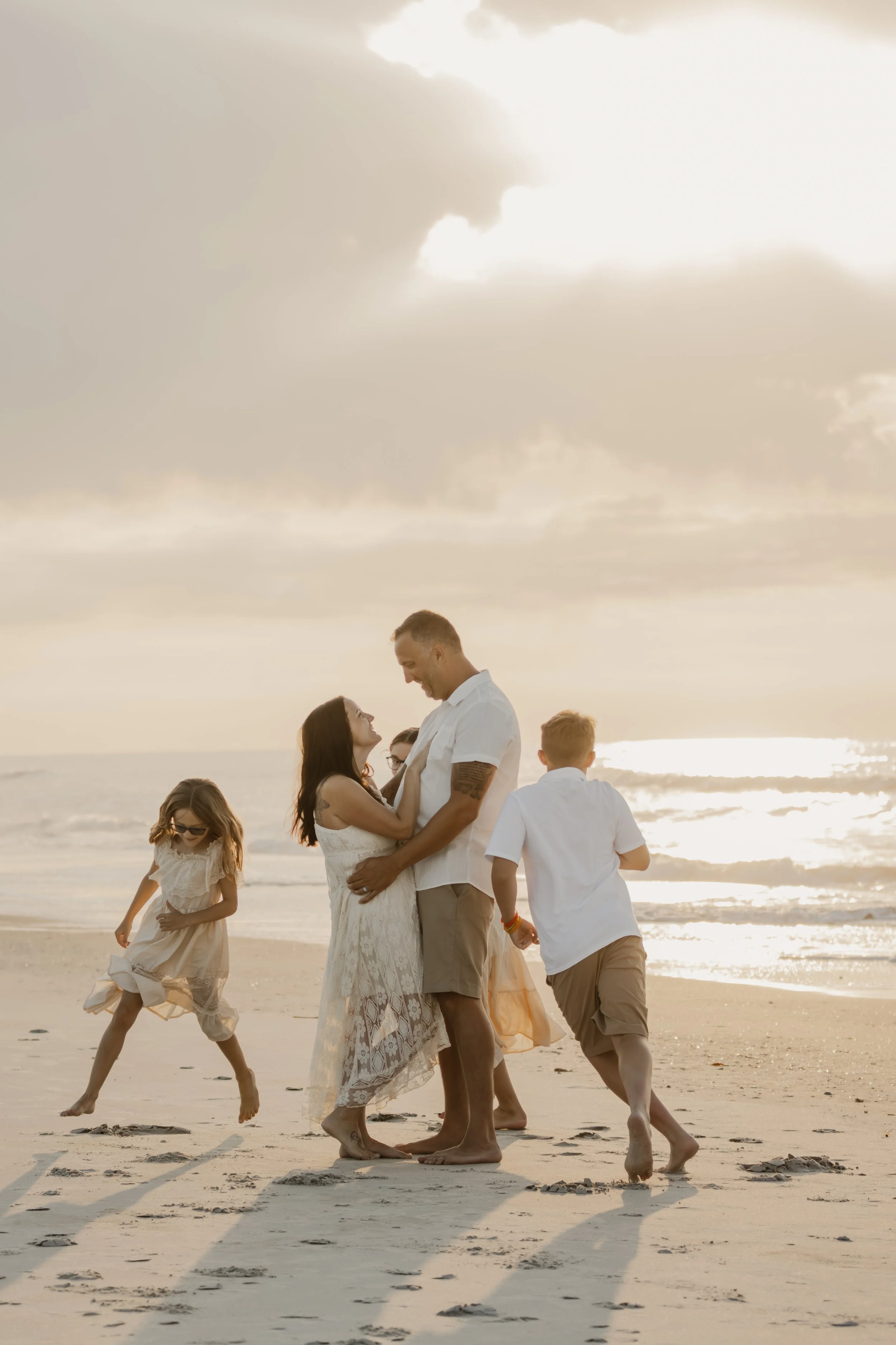 A group of people, including children and adults, enjoying a sunset on the beach, with a man and woman embracing in the center.