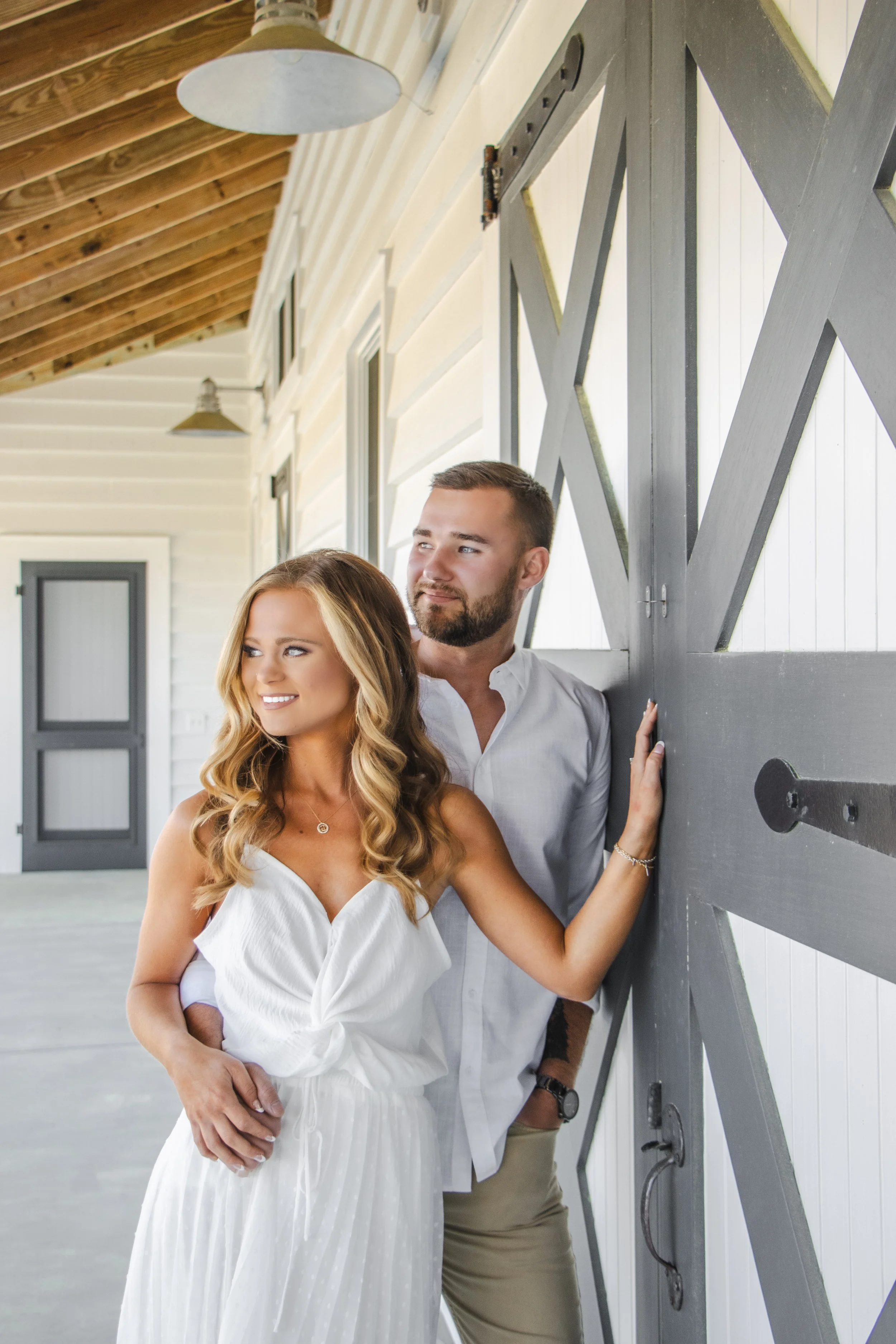 A couple standing inside a modern barn, with the woman wearing a white dress and the man in a white shirt, leaning against a large black sliding barn door.