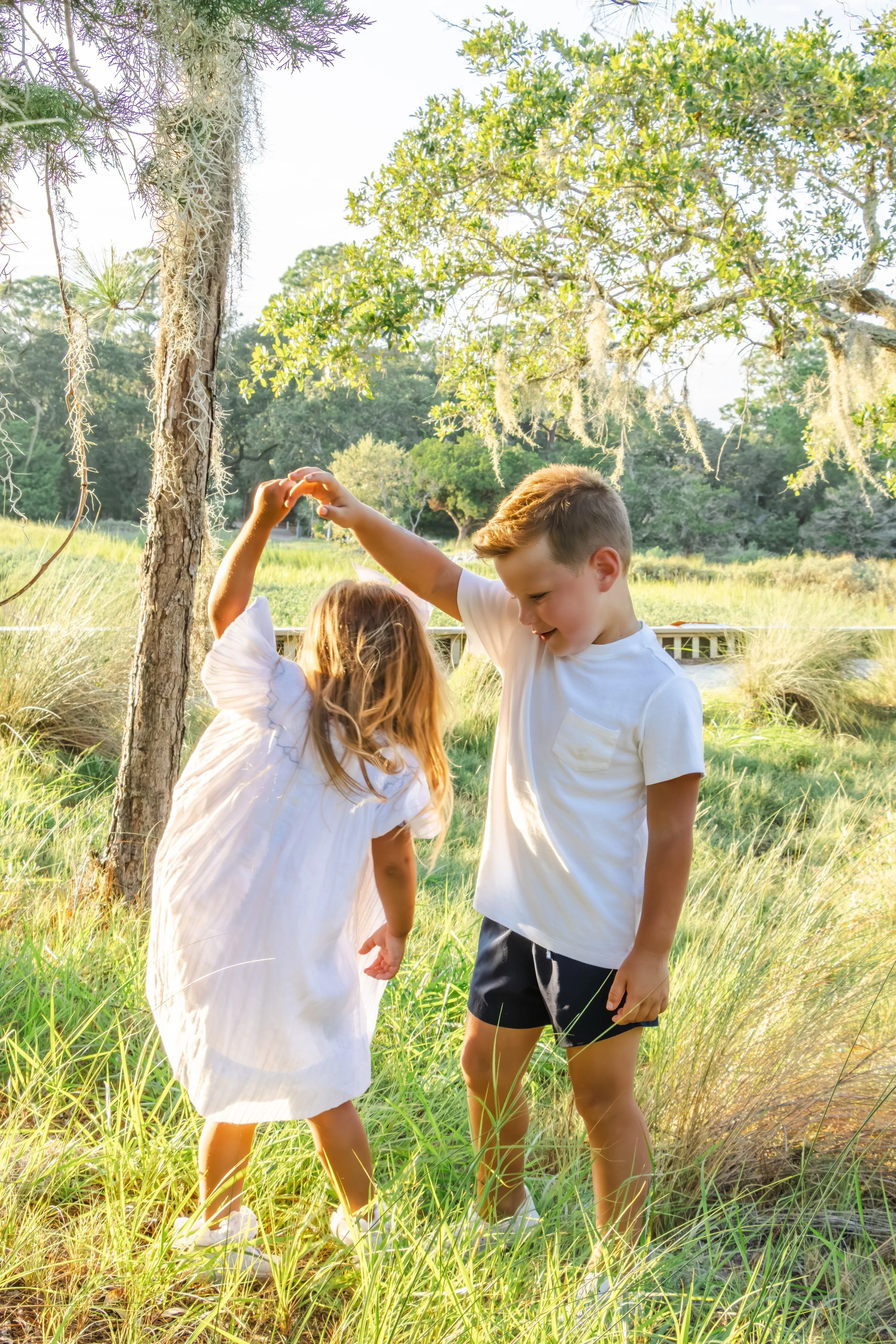 A boy and girl dancing together in a grassy outdoor area surrounded by trees on a sunny day.