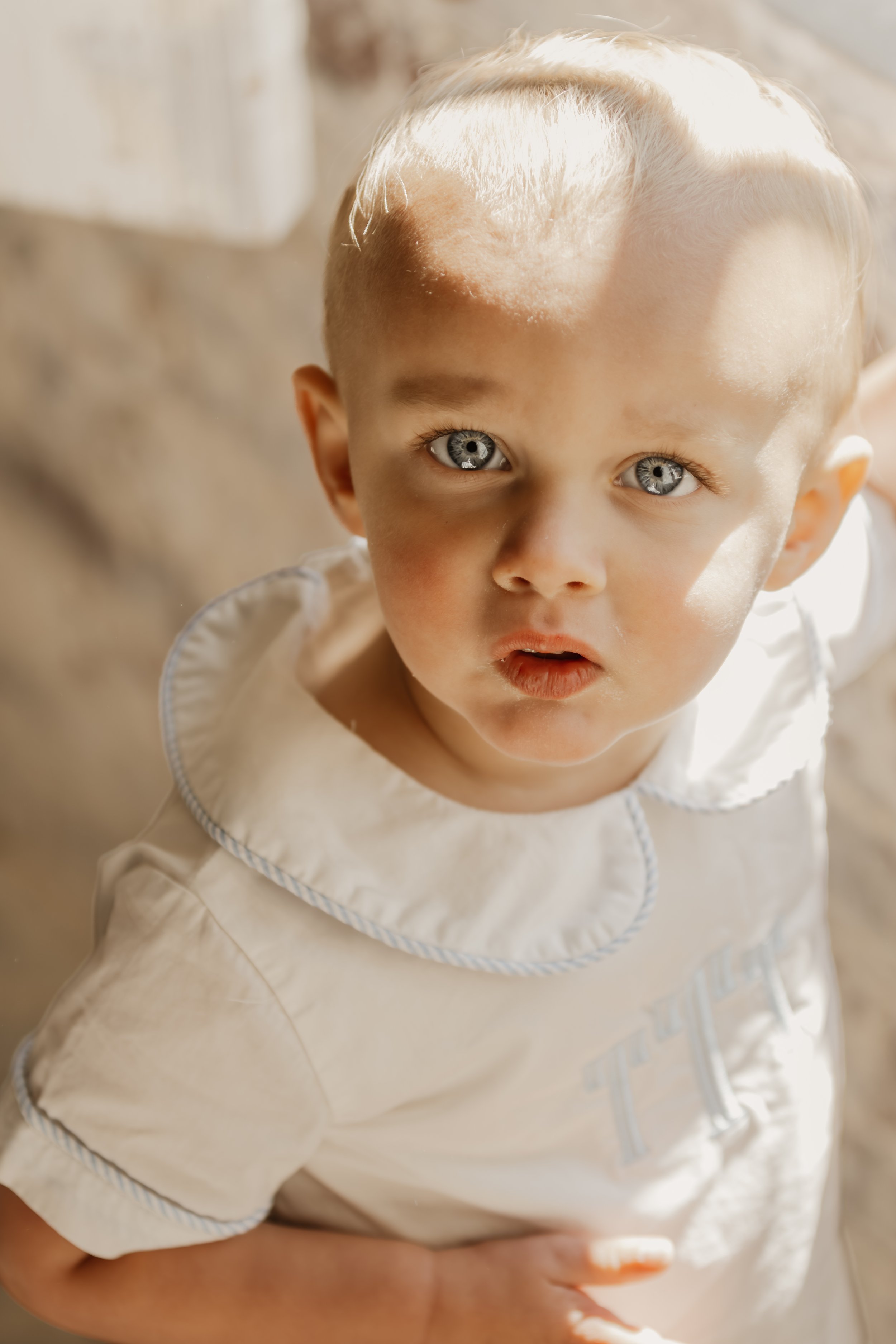 A young boy with blonde hair and blue eyes looking up at the camera with a curious expression, wearing a cream-colored shirt with a collar.