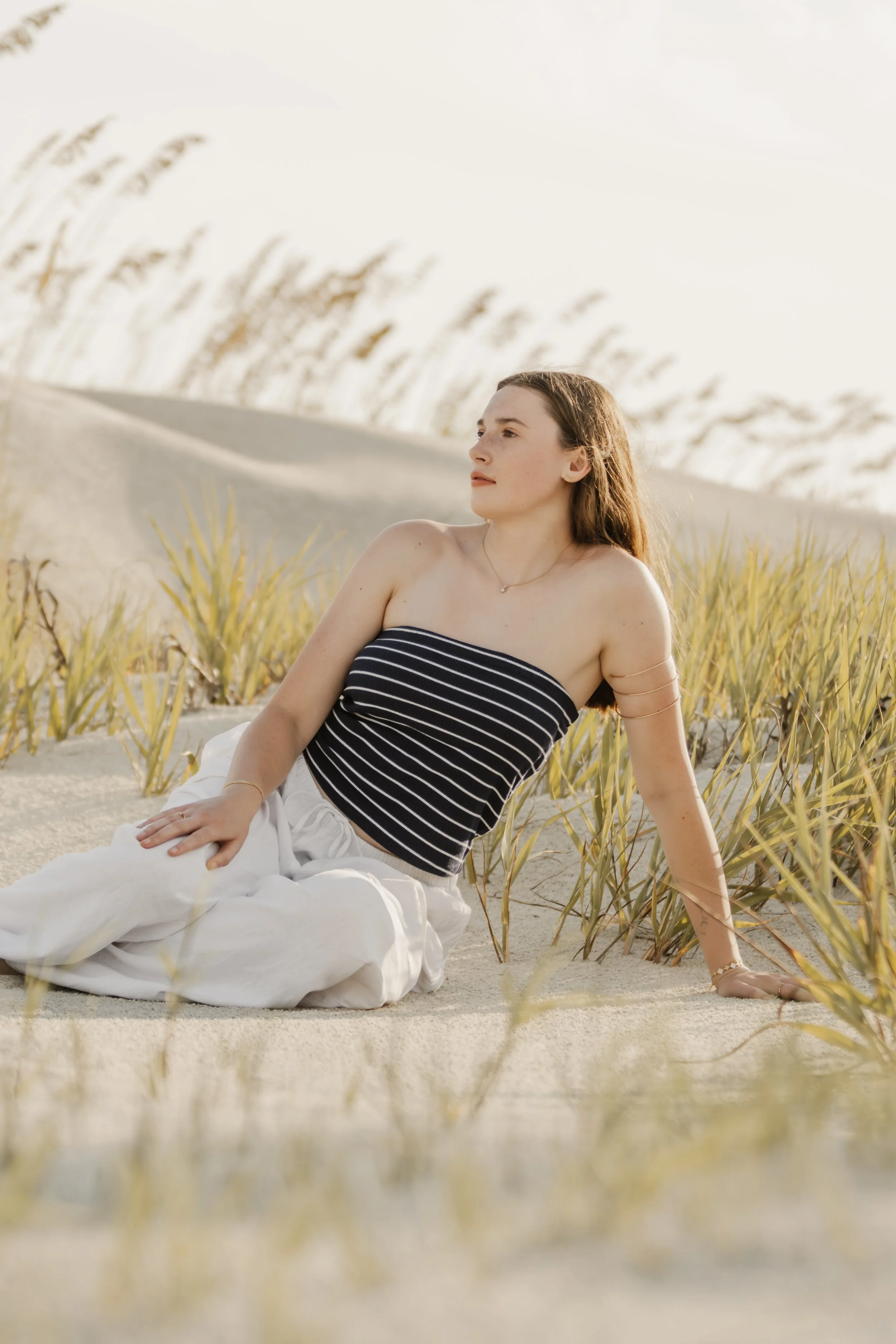 A young woman in a strapless black and white striped top and white pants, sitting on a sandy beach with tall grass and sand dunes in the background, looking into the distance.