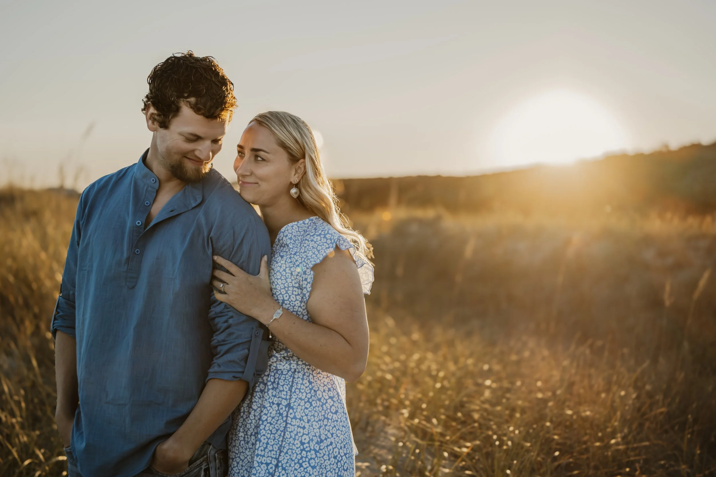 A couple standing close together outdoors during sunset, embracing and smiling at each other in a field of tall grass.