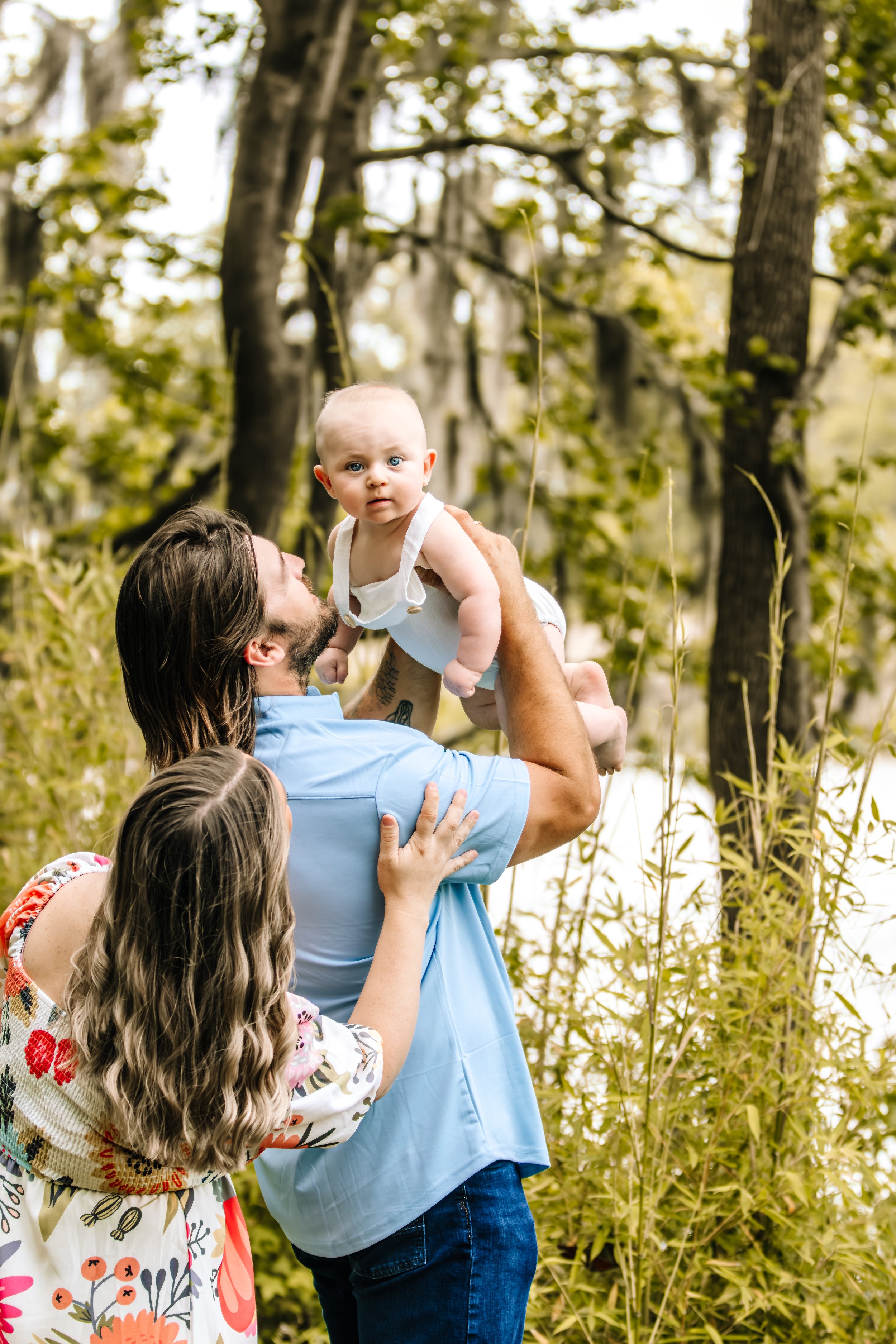 A man with long hair and a beard holding a baby girl in an outdoor wooded area, with a woman beside them touching the man as he lifts the baby. The scene is surrounded by trees and foliage.