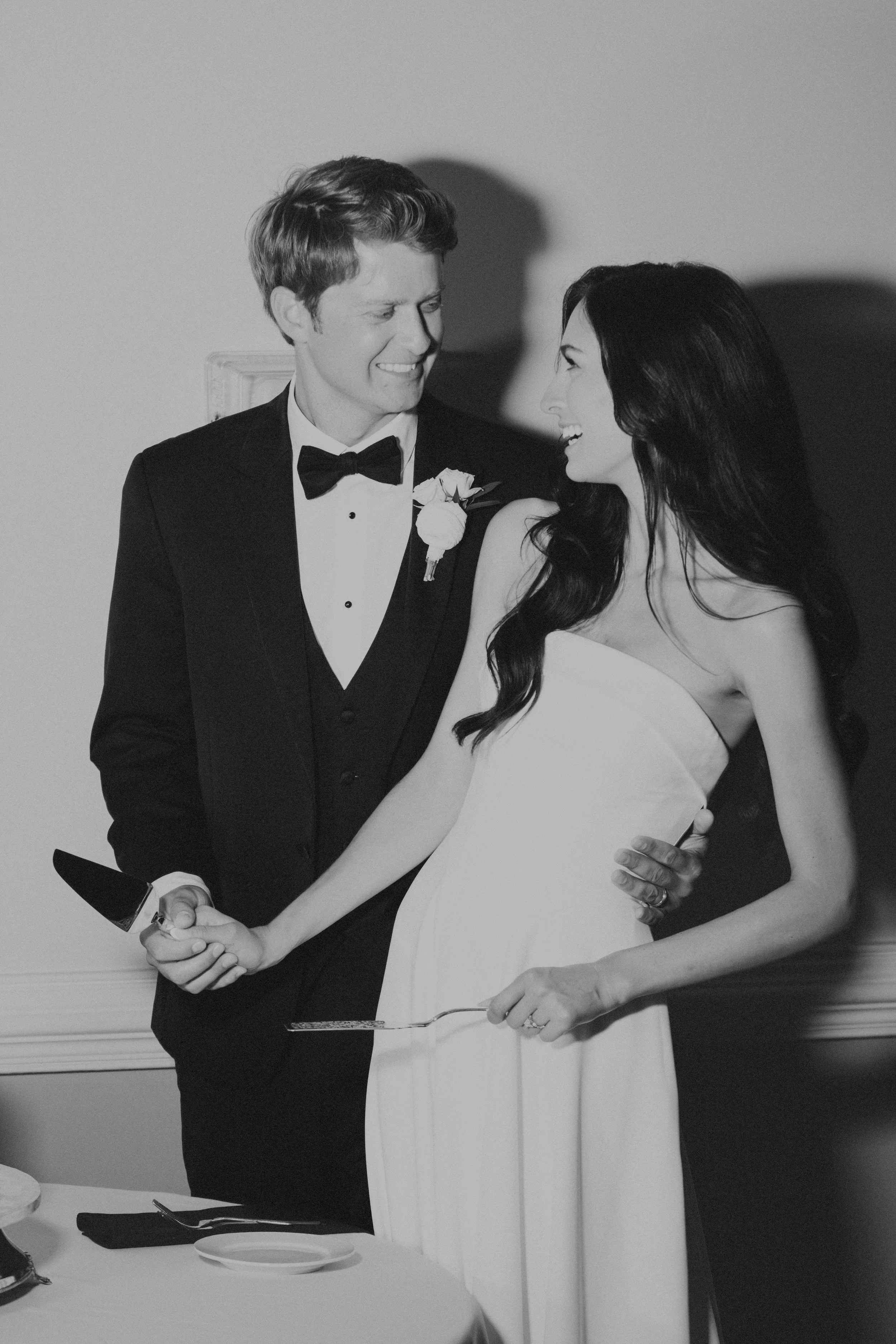 A newlywed couple dressed in wedding attire, smiling and holding a cake serving knife together at their wedding reception.