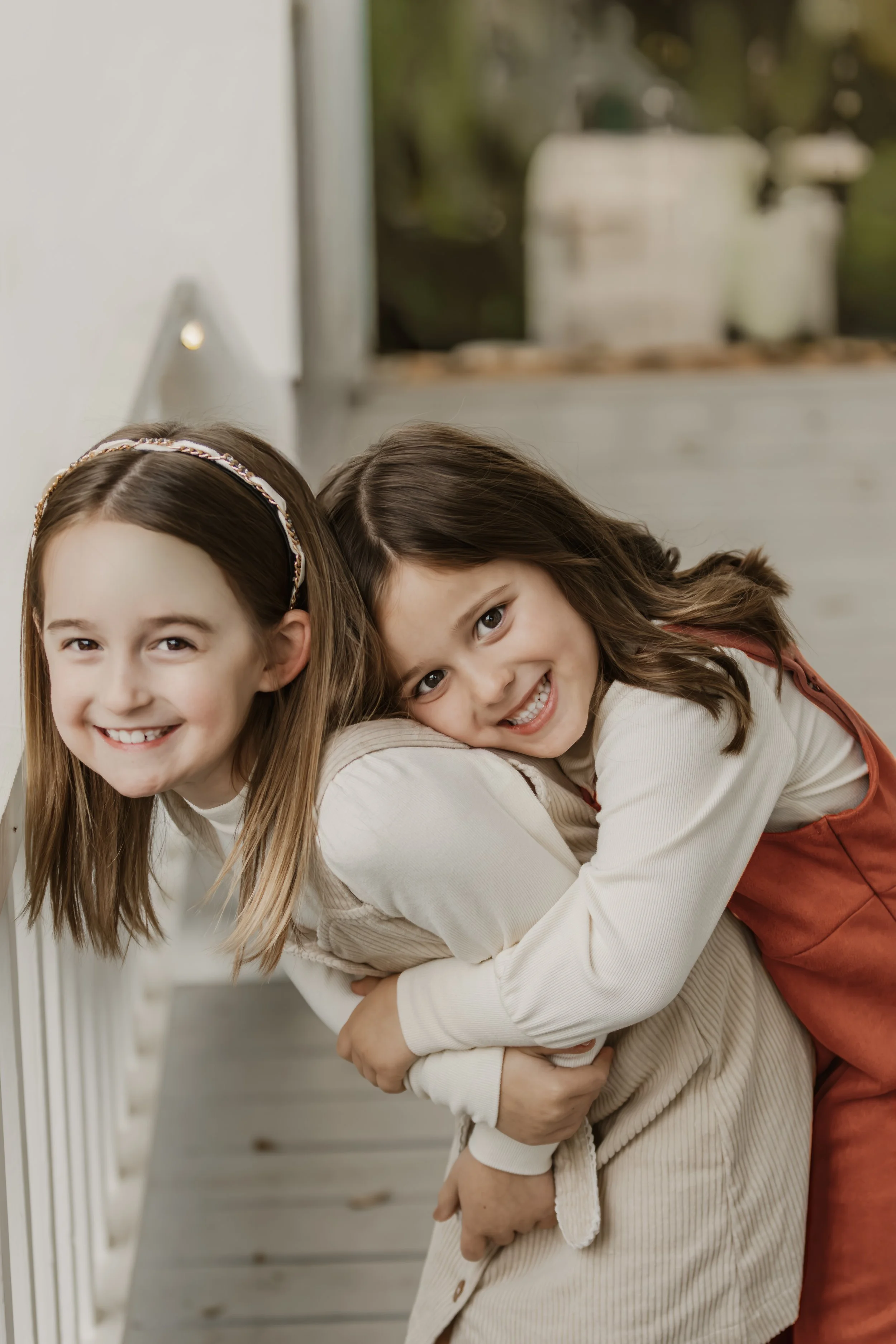 Two young girls smiling, one embracing the other from behind, in a cozy indoor setting with a window and plants in the background.